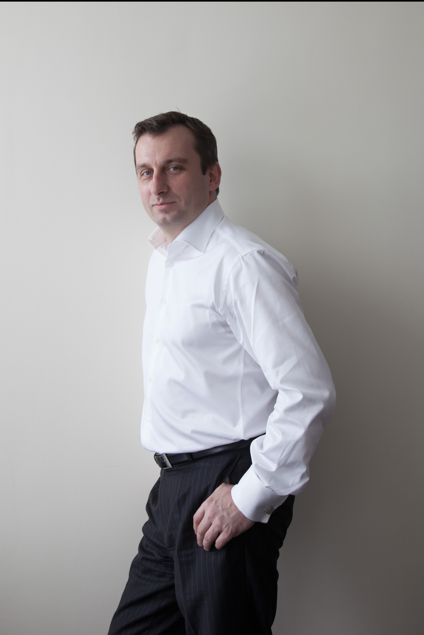 Mężczyzna w białej koszuli i ciemnych spodniach w paski, pozuje na jasnym tle. Widoczny profil twarzy i część sylwetki. Elegancki strój, biznesowy charakter.