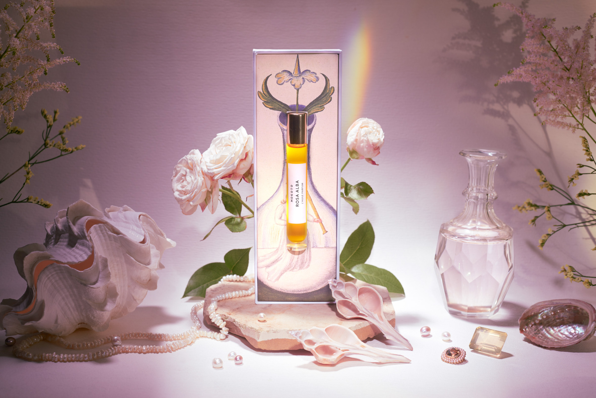 Artystyczna fotografia produktowa: flakon perfum Mokoto Rosa Alba na kamiennej podstawce, otoczony kwiatami, muszlami i perłami w pastelowej kolorystyce z efektem tęczy.