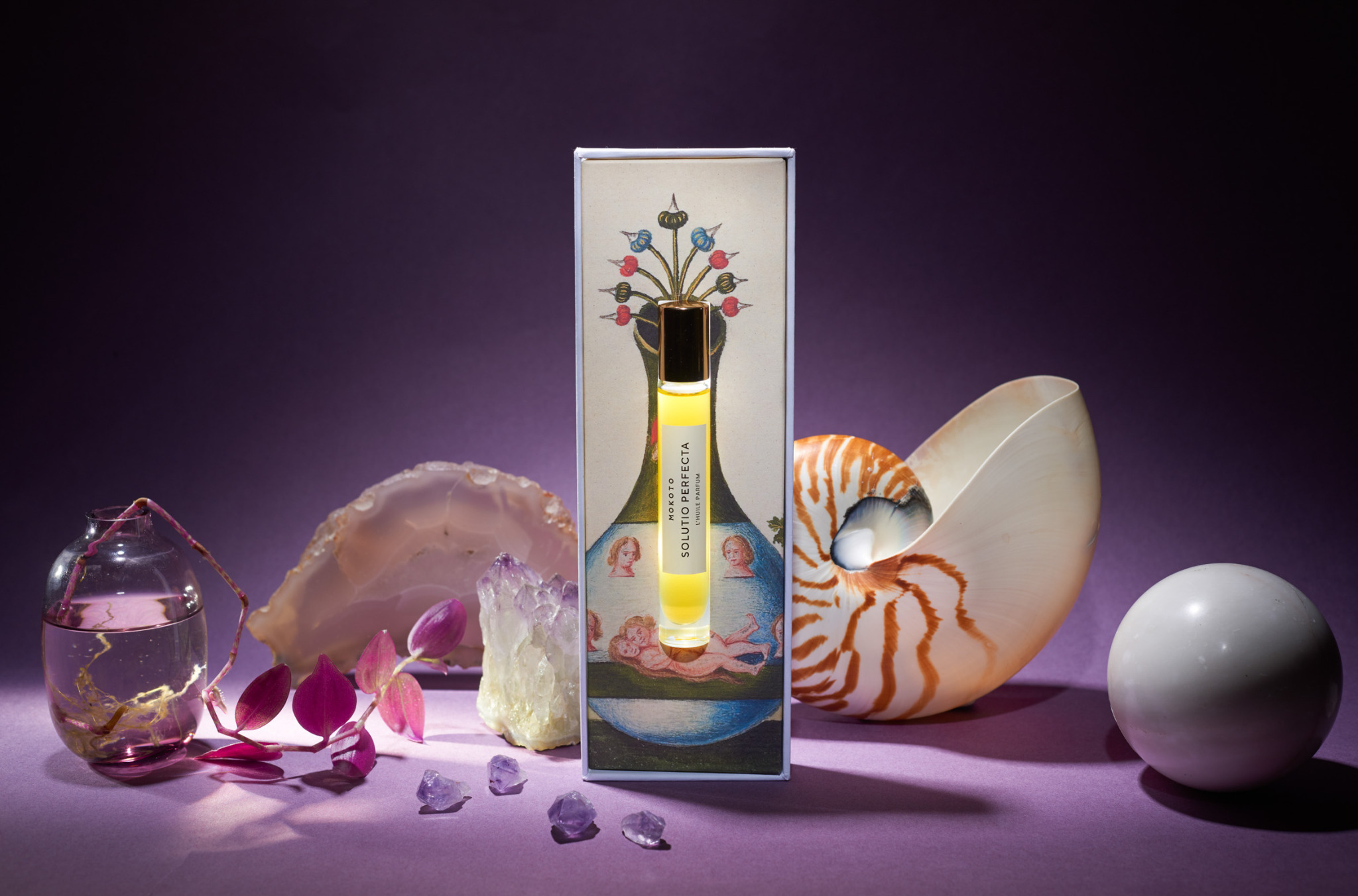 Zdjęcie flakonika perfum Mokoto Solutio Perfecta w ozdobnym pudełku, otoczonego kryształami, muszlą i szklanym wazonem na fioletowym tle. Produkt oświetlony z góry.