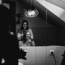 Maja Kasztelan fotograf - Czarno-białe zdjęcie zrobione w łazience, przedstawia dwie uśmiechnięte kobiety; jedna siedzi z włosami częściowo upiętymi, trzymając szklankę, druga stoi za nią i poprawia jej włosy; widoczne...