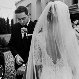 Maja Kasztelan fotograf - Czarno-białe zdjęcie nowożeńców: pan młody w smokingu sprawdza godzinę na zegarku, panna młoda w koronkowej sukni i welonie trzyma bukiet ślubny, obok czarny samochód.