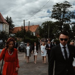 Maja Kasztelan fotograf - Grupa elegancko ubranych gości weselnych idzie drogą w kierunku kościoła, w tle widoczne budynki mieszkalne i pochmurne niebo.