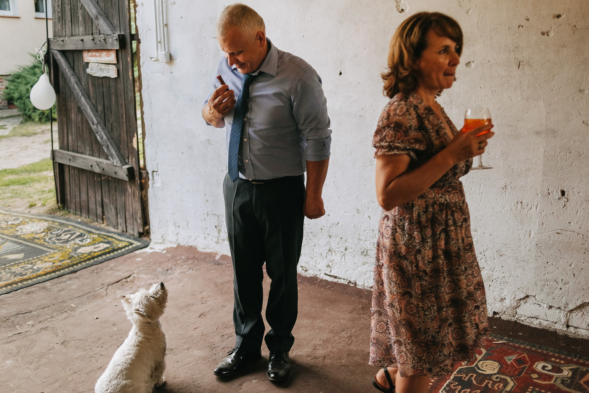 Mężczyzna w koszuli i krawacie poprawia szminkę przed kobietą z drinkiem i psem rasy West Highland White Terrier na tle rustykalnych drzwi i ściany.