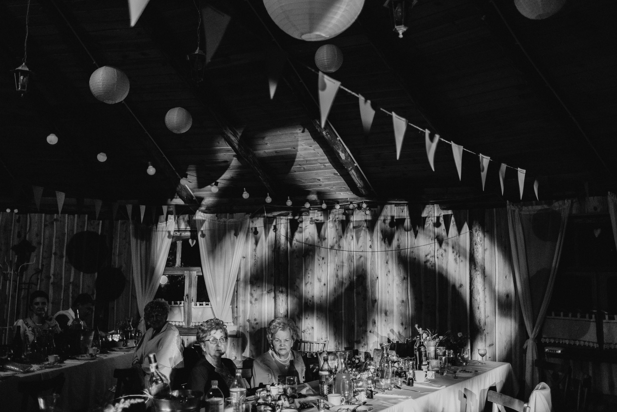 Czarno-białe zdjęcie sali z drewnianymi ścianami i dekoracjami w postaci papierowych lampionów oraz girland z trójkątnych chorągiewek. Goście siedzą przy długim stole nakrytym białym obrusem...
