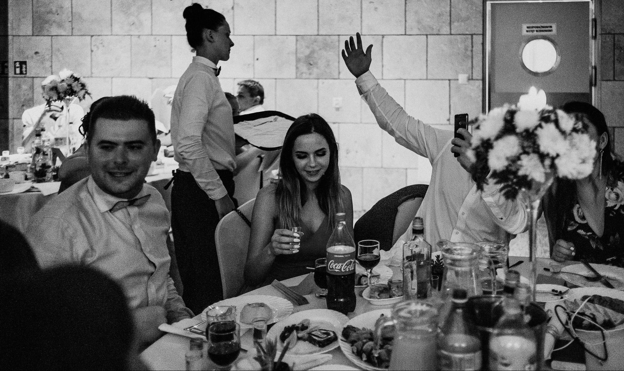 Czarno-białe zdjęcie z przyjęcia: uśmiechnięty mężczyzna w muszce, kobieta z kieliszkiem w dłoni, kelner w tle, podniesiona ręka osoby robiącej zdjęcie telefonem, butelka Coca-Coli na stole.