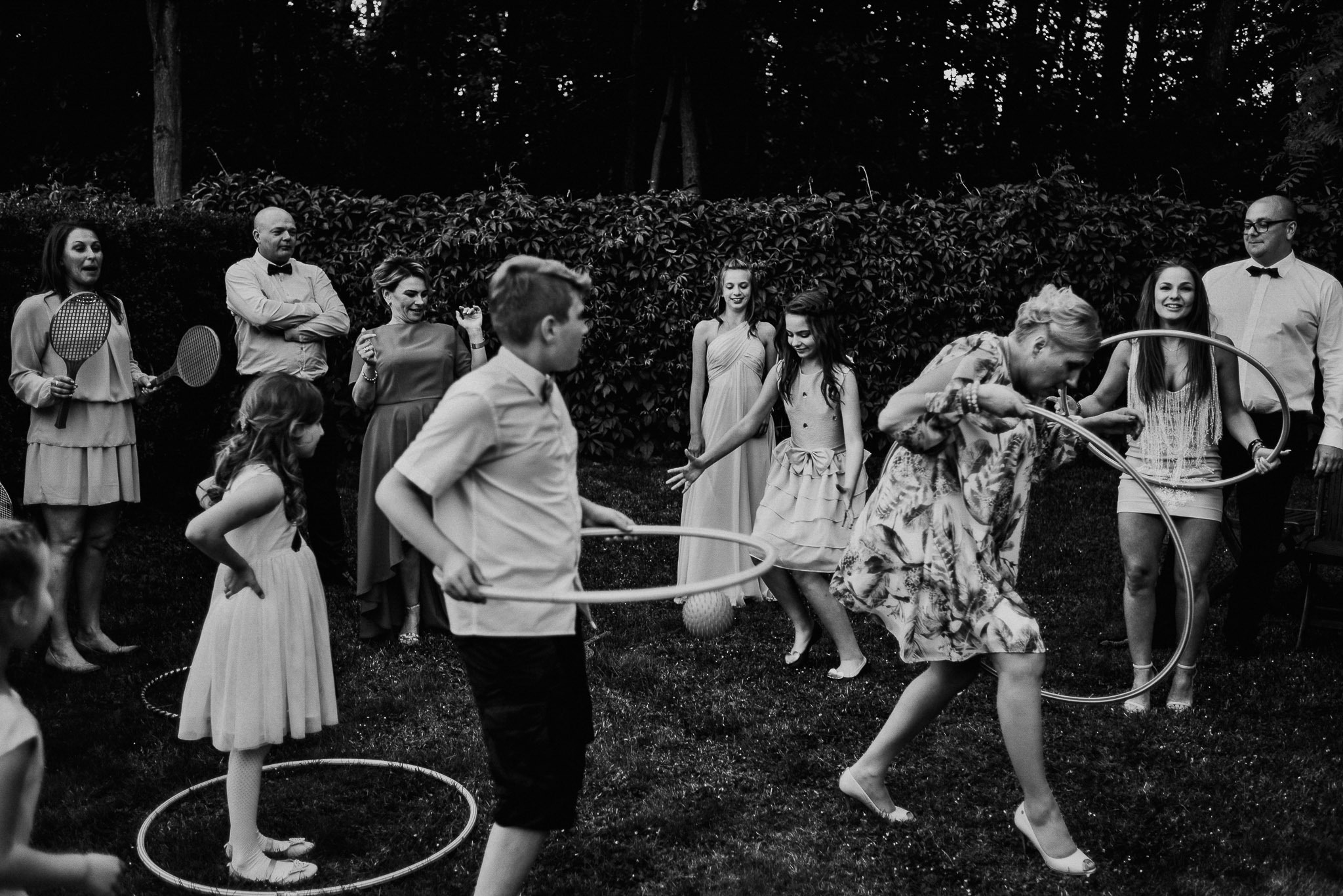 Czarno-białe zdjęcie przedstawia grupę osób na trawniku podczas zabawy hula-hoop, w tle widoczny żywopłot, a niektórzy trzymają rakiety tenisowe.