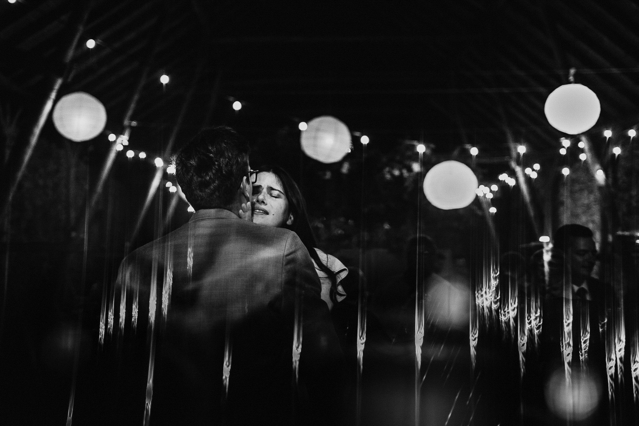 Czarno-białe zdjęcie pary obejmującej się na tle dekoracji świetlnych, widoczne odbicia w szklanej powierzchni.