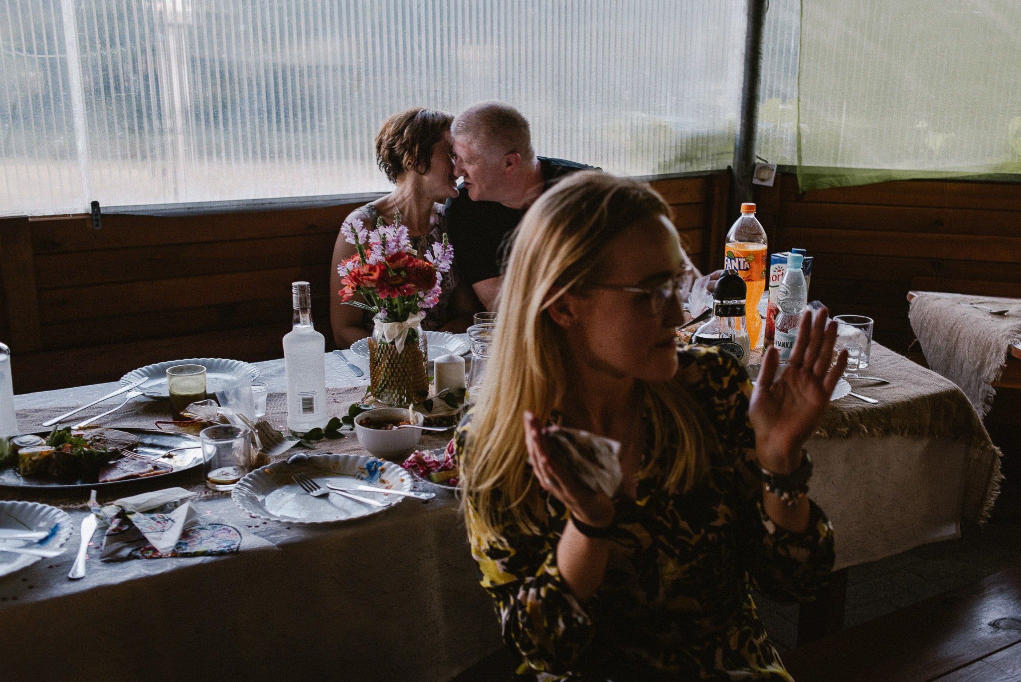 Para całująca się w tle, widoczna znad stołu z resztkami jedzenia i bukietem kwiatów, na pierwszym planie kobieta z uniesionymi rękami w żółto-czarnej bluzce.