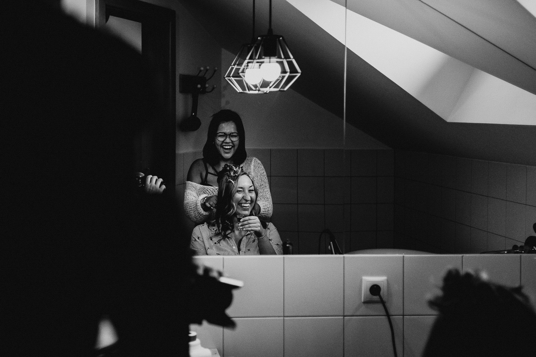 Czarno-białe zdjęcie zrobione w łazience, przedstawia dwie uśmiechnięte kobiety; jedna siedzi z włosami częściowo upiętymi, trzymając szklankę, druga stoi za nią i poprawia jej włosy; widoczne...