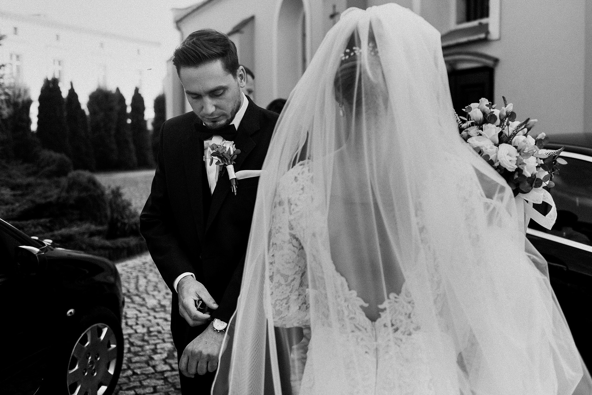 Czarno-białe zdjęcie nowożeńców: pan młody w smokingu sprawdza godzinę na zegarku, panna młoda w koronkowej sukni i welonie trzyma bukiet ślubny, obok czarny samochód.