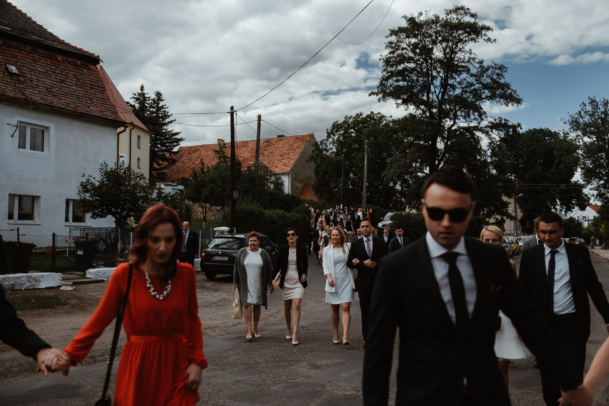 Grupa elegancko ubranych gości weselnych idzie drogą w kierunku kościoła, w tle widoczne budynki mieszkalne i pochmurne niebo.