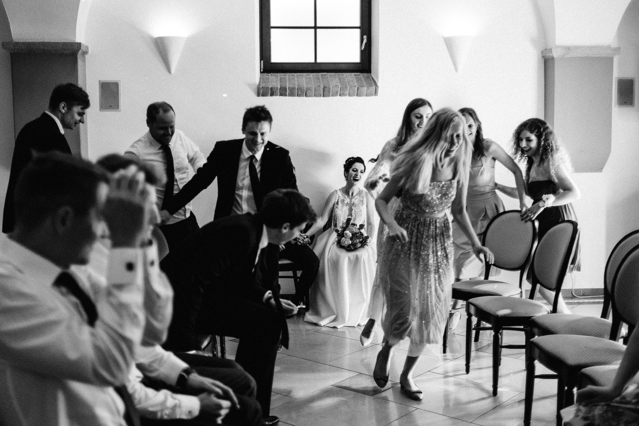 Czarno-białe zdjęcie z wesela: panna młoda siedzi na krześle z bukietem, wokół niej tańczą goście, w tle okno.