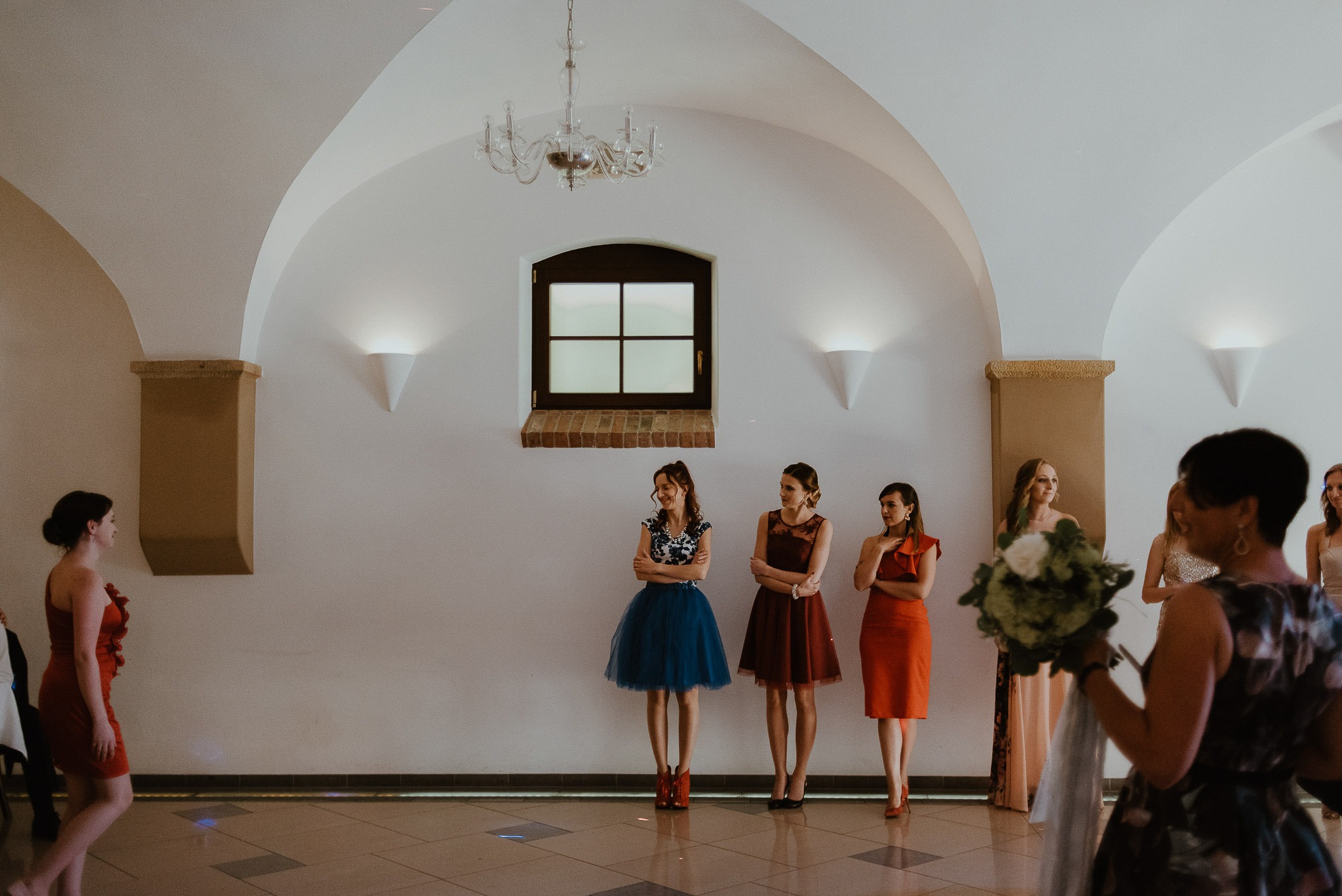 Panna młoda z bukietem i druhny w kolorowych sukienkach w przestronnym, jasnym wnętrzu z łukowym sufitem i żyrandolem.