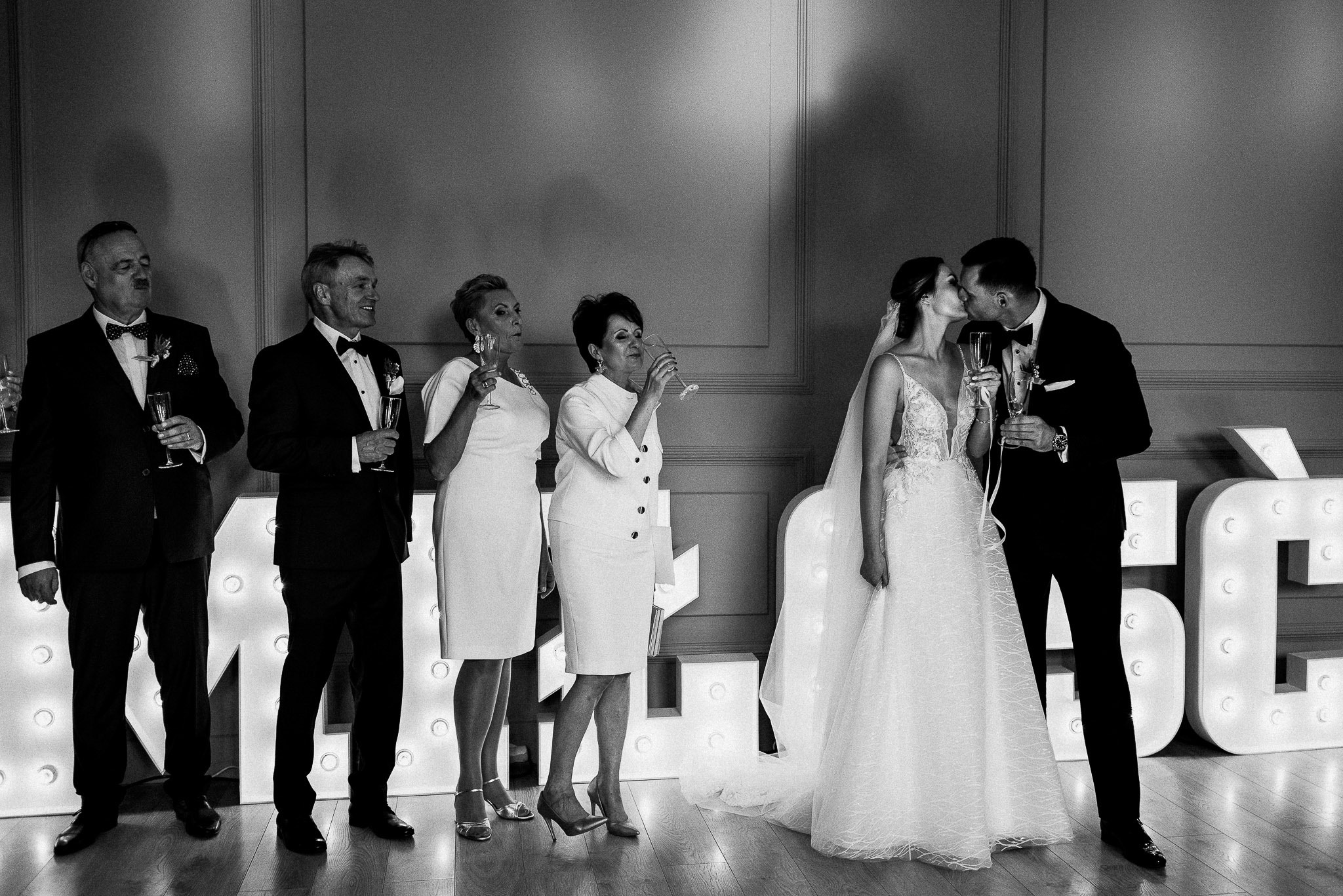 Czarno-białe zdjęcie nowożeńców całujących się, trzymających kieliszki szampana, z gośćmi weselnymi i podświetlanymi literami w tle.