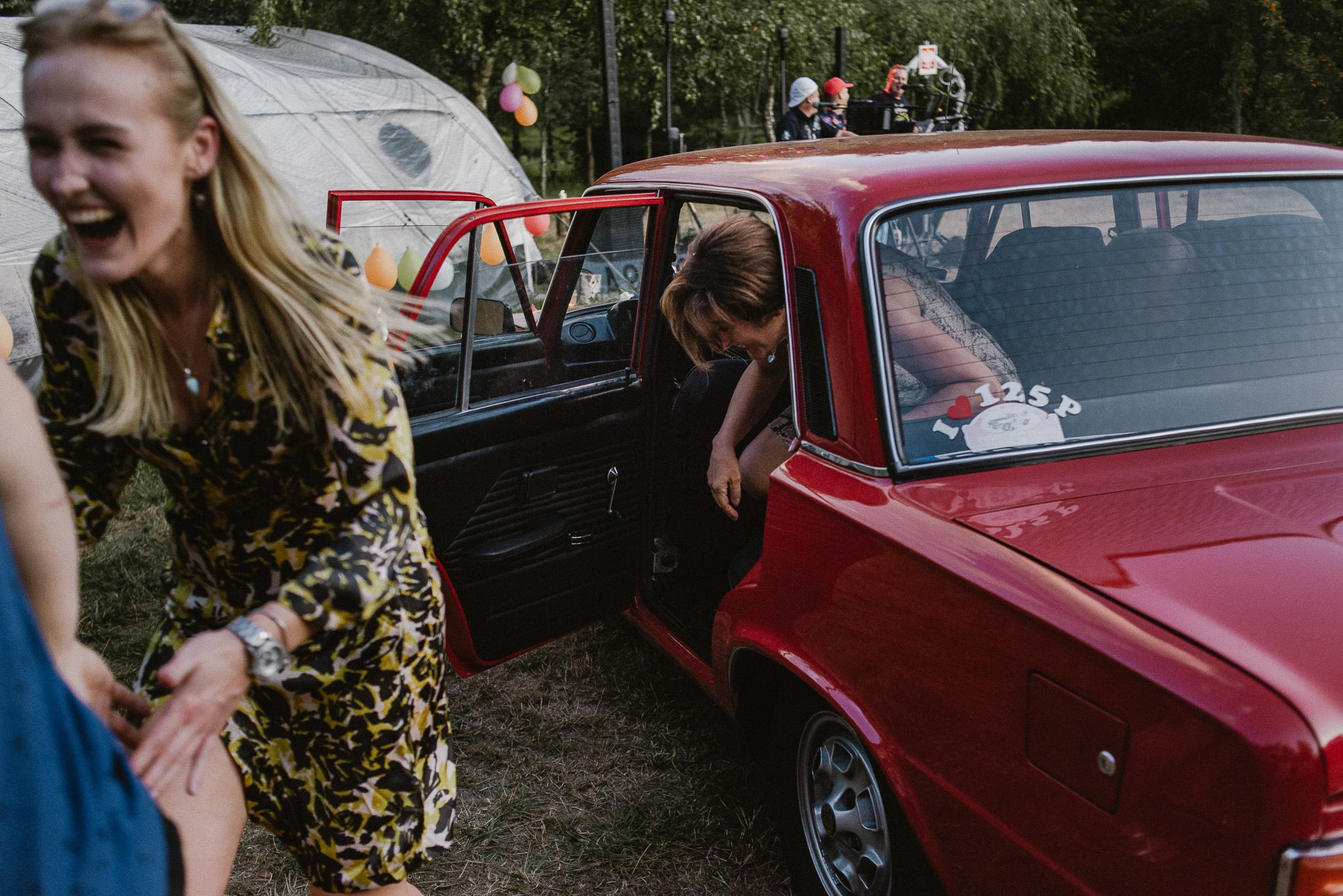 Uśmiechnięta blondynka w kwiecistej sukience, wychodząca z czerwonego samochodu z naklejką 'I love 125p', w tle namiot i balony, impreza plenerowa.