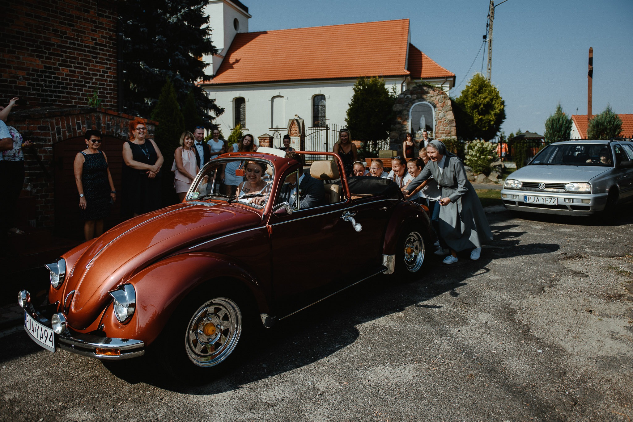 Panna młoda uśmiechnięta siedzi za kierownicą brązowego kabrioletu VW Garbus, w tle goście weselni i siostra zakonna pomagająca w przepychaniu auta.