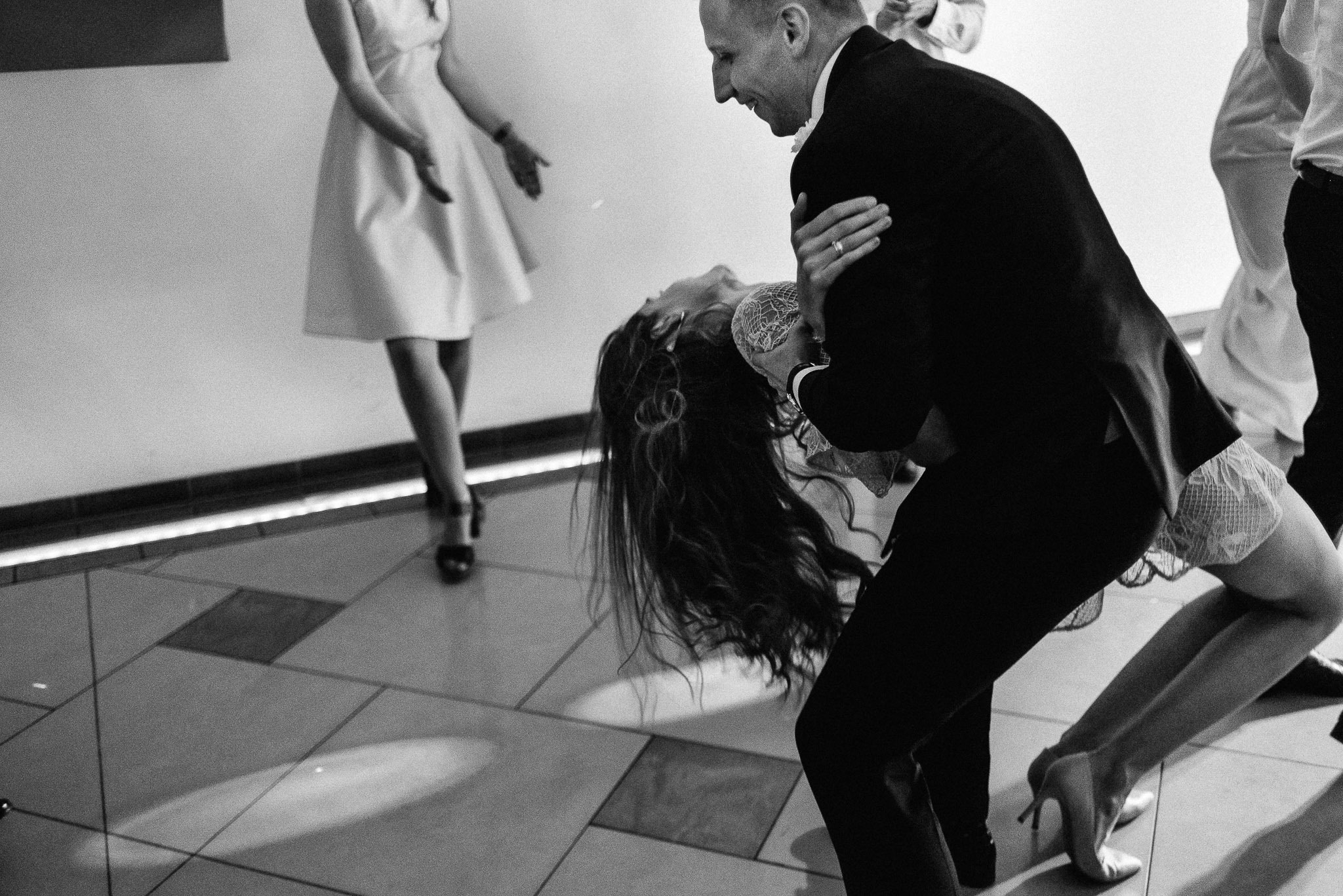 Para w tańcu podczas uroczystości, mężczyzna w czarnym garniturze trzyma kobietę w jasnej sukni, przechyloną do tyłu, w tle widoczni inni goście.