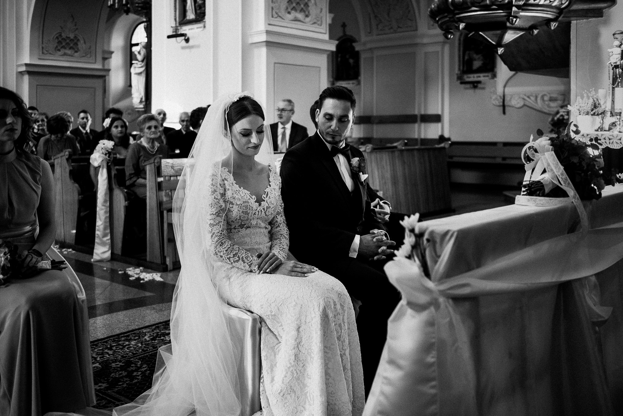 Czarno-białe ujęcie pary młodej siedzącej w kościele w Gdańsku podczas ceremonii ślubnej, z gośćmi w tle i dekoracjami kwiatowymi na stole przed nimi.