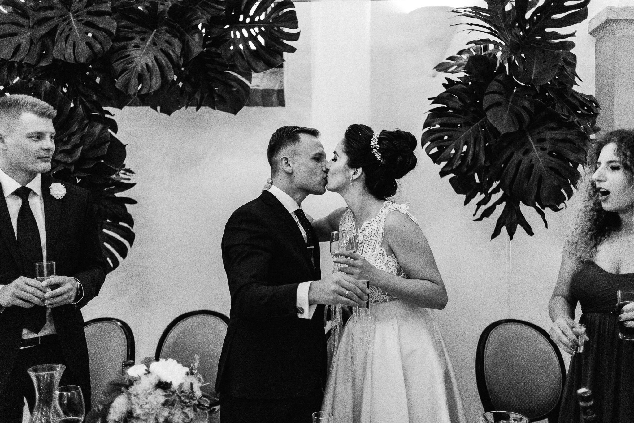 Czarno-białe zdjęcie całującej się pary młodej na tle dekoracji z liści monstery, goście weselni w tle trzymają kieliszki.