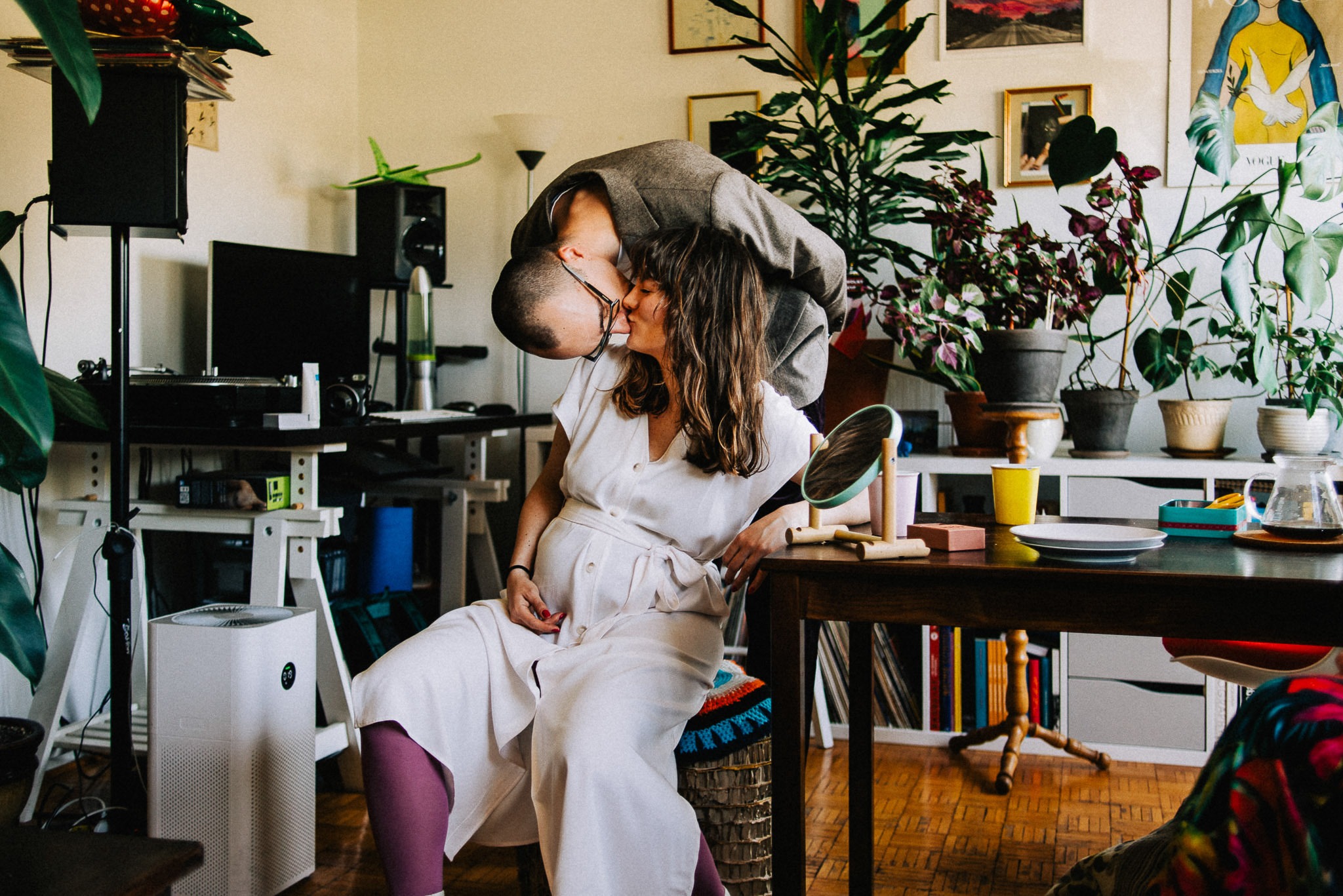 Czułe zbliżenie kobiety w ciąży siedzącej na krześle, całowanej przez mężczyznę w szarej marynarce, w tle pokój z roślinami, biurkiem i dekoracjami.