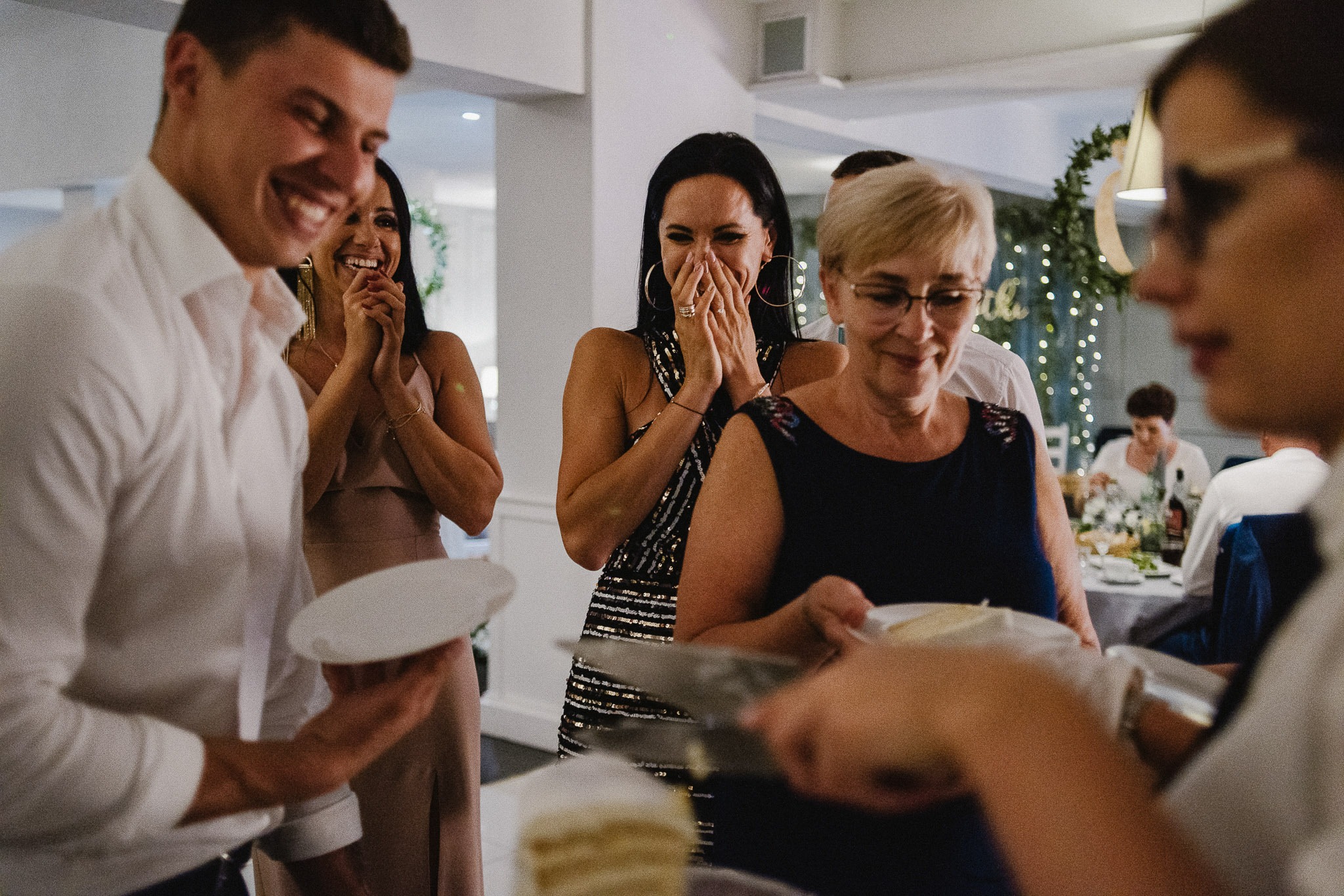 Goście weselni z uśmiechem obserwują krojenie tortu, widoczna kobieta w cekinowej sukience zakrywająca usta dłońmi, w tle dekoracje świetlne.