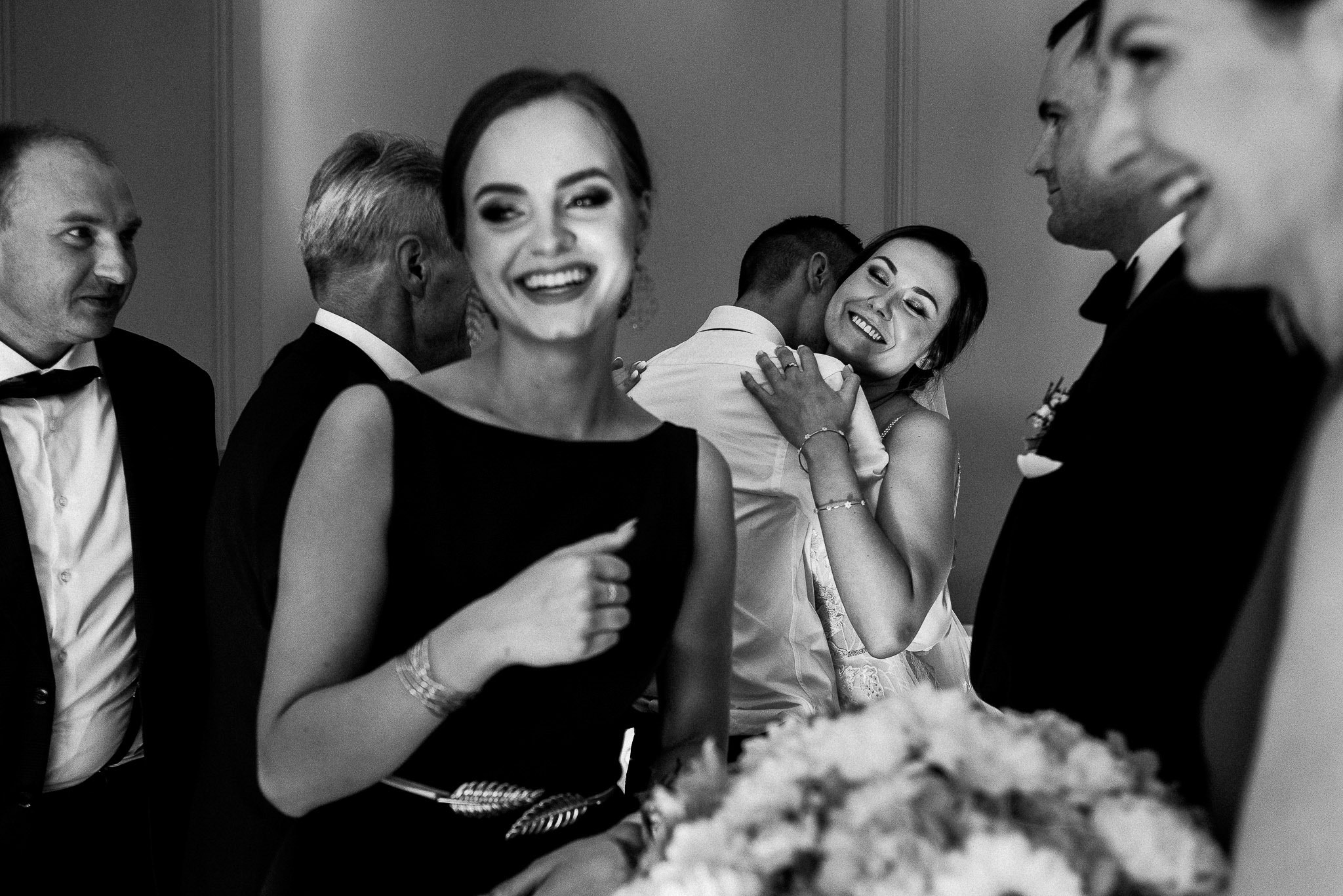 Czarno-białe zdjęcie z wesela, uśmiechnięta kobieta w czarnej sukience z ozdobnym paskiem na pierwszym planie, w tle para młoda w objęciach i goście weselni.