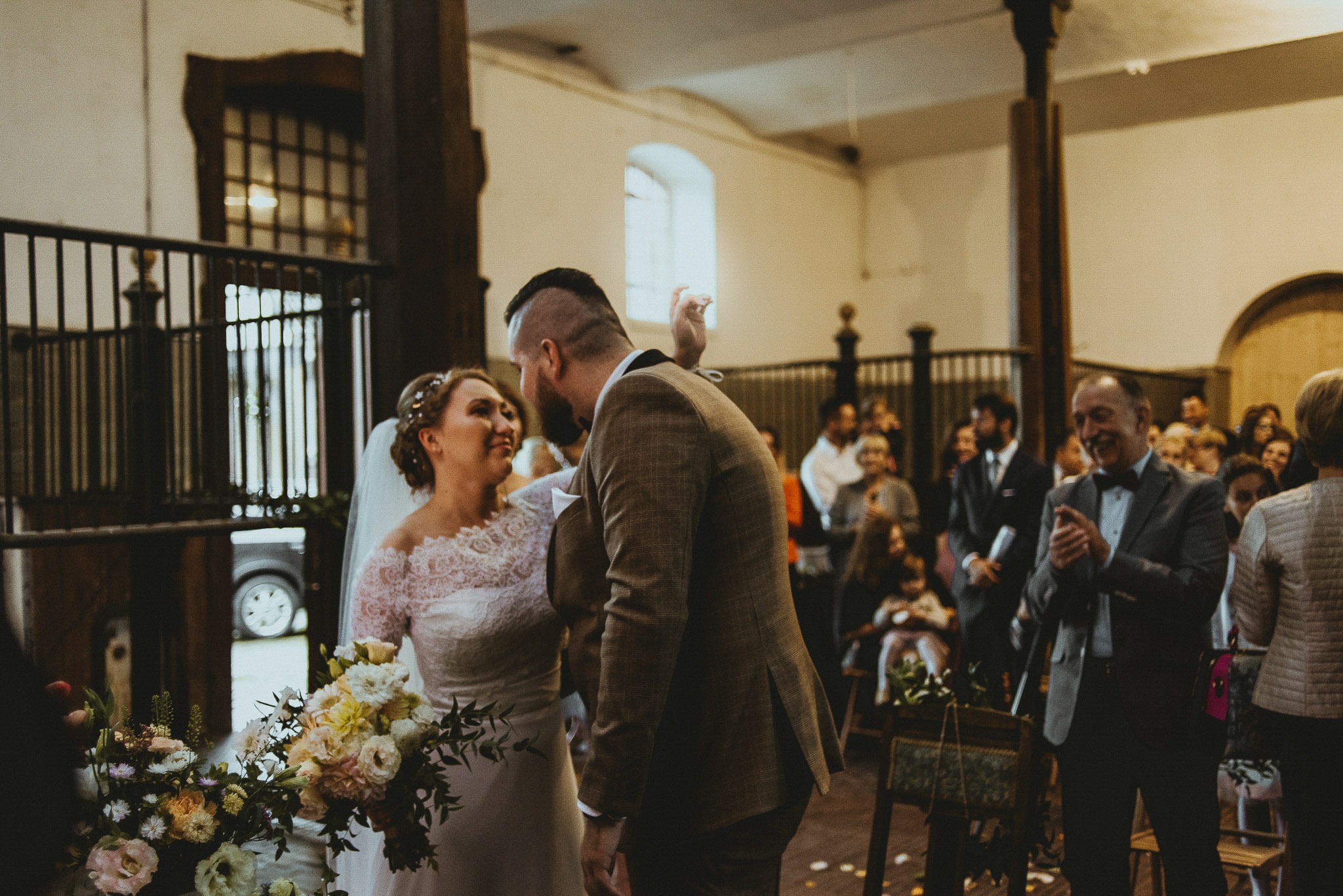Panna młoda w koronkowej sukni spogląda z uśmiechem na pana młodego w brązowym garniturze podczas ceremonii ślubnej w przestronnym wnętrzu z drewnianymi kolumnami i gośćmi w tle.