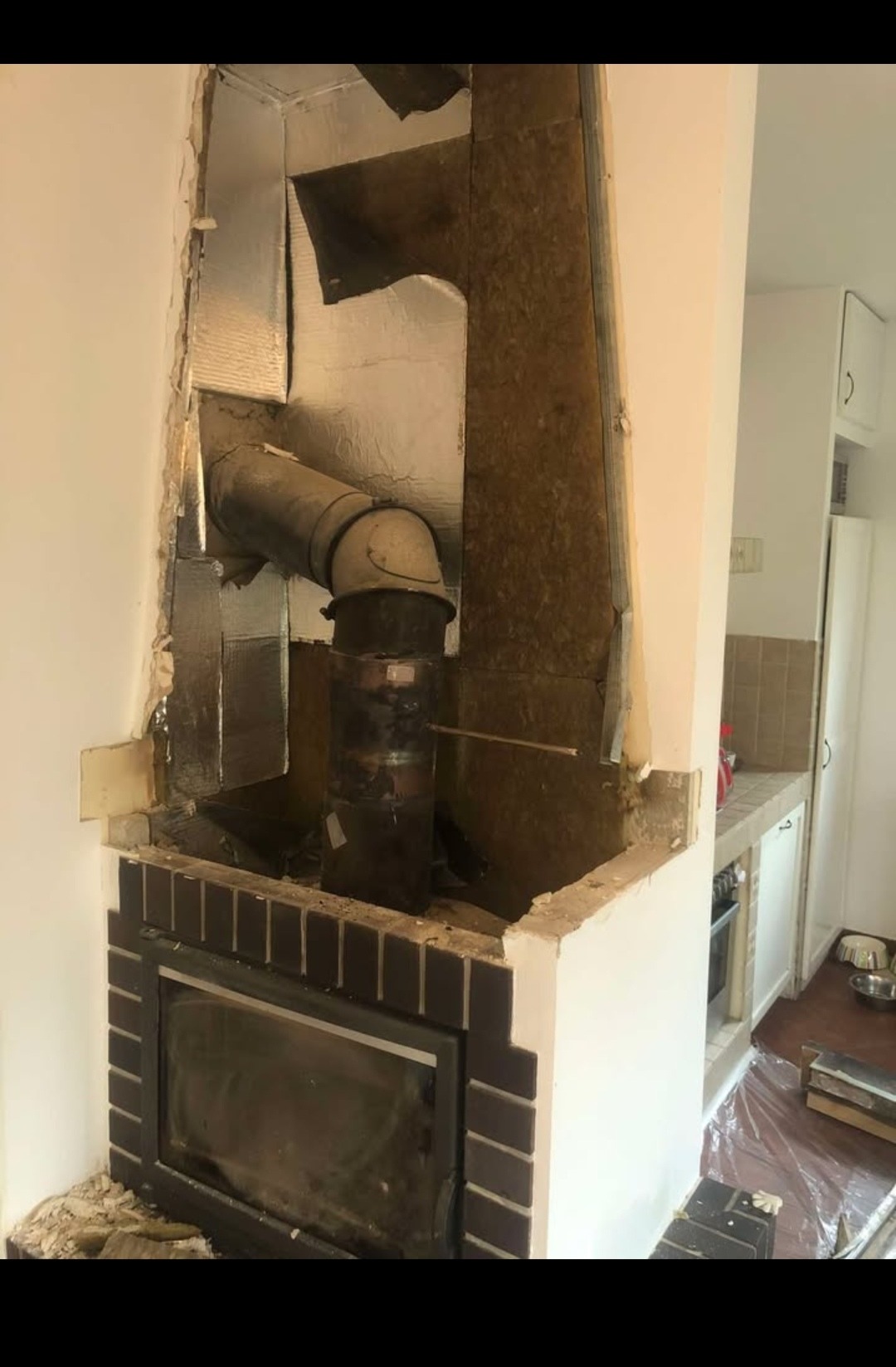 Odsłonięty komin z metalową rurą w trakcie prac remontowych, widoczne płyty izolacyjne i uszkodzone ściany wokół obudowy kominka z ciemnej cegły.