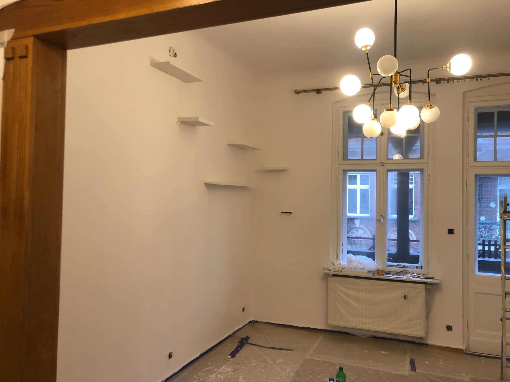 Świeżo pomalowane na biało wnętrze pokoju z elementami przygotowanymi do montażu półek, widoczne zabezpieczenia podłogi folią malarską i grzejnika, charakterystyczny żyrandol z kulistymi kloszami.