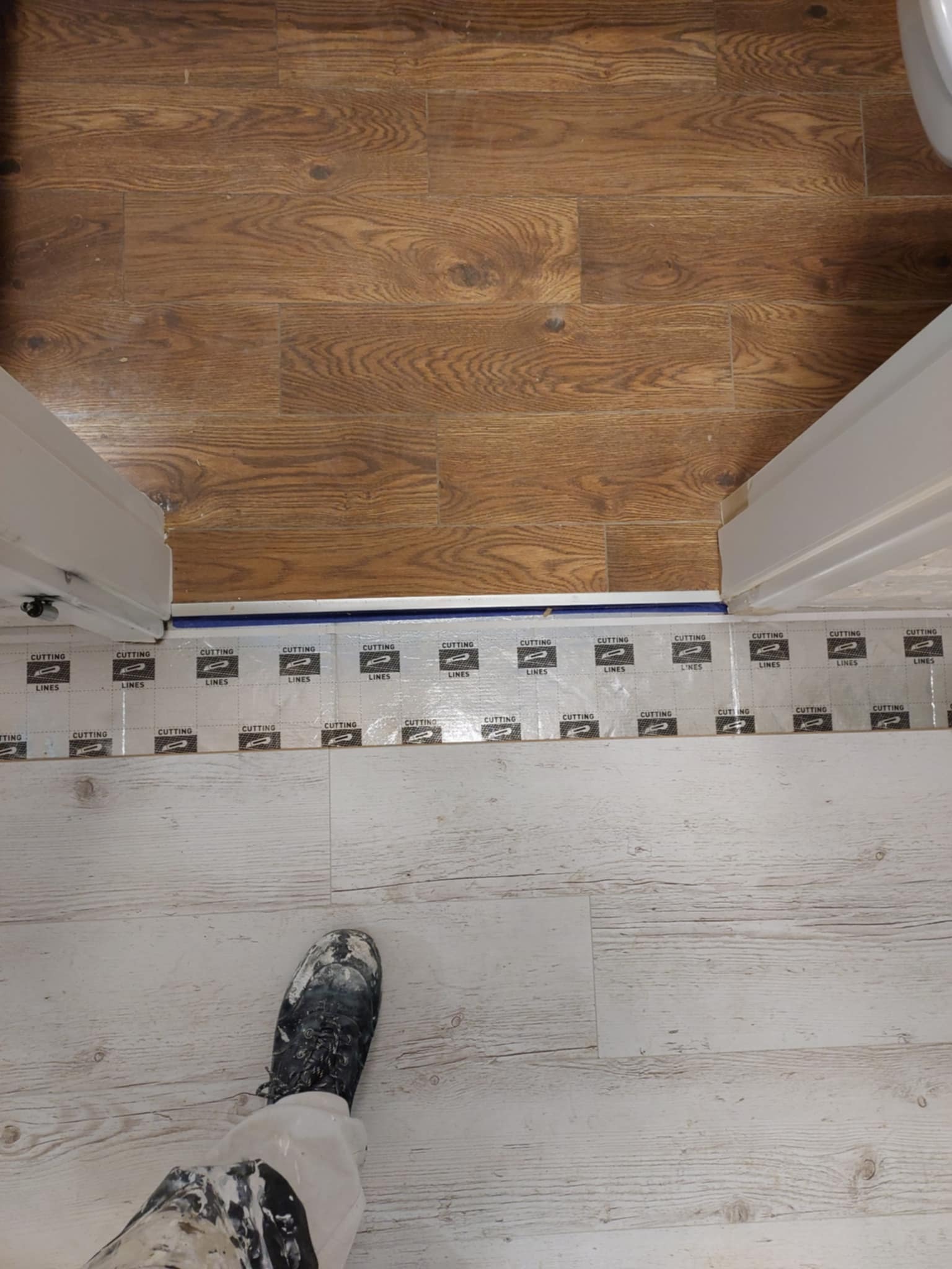 Ochrona podłogi przy użyciu folii z napisem 'CUTTING LINES' podczas prac wykończeniowych, widoczny fragment spodni roboczych z zabrudzeniami farby i but roboczy.