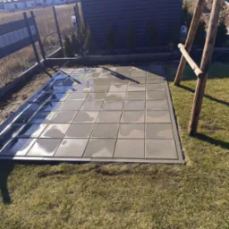 Świeżo ułożona, mokra kostka brukowa na posesji, z widocznym fragmentem ogrodzenia i drewnianej konstrukcji huśtawki w tle, w słoneczny dzień.