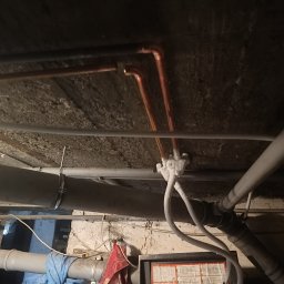 W.R. REMONTY WALDEMAR RATAJCZAK - Instalacja rur miedzianych i PCV pod stropem, widoczne łączenia i mocowania. Ujęcie od dołu, ciemne tło uwydatnia detale instalacji.