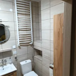 Nowoczesna łazienka z beżowymi płytkami, białym grzejnikiem drabinkowym i okrągłym lustrem z podświetleniem. Widoczna toaleta i umywalka z szafką w kolorze drewna.