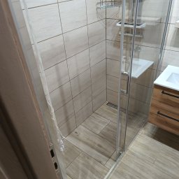 W.R. REMONTY WALDEMAR RATAJCZAK - Nowoczesna kabina prysznicowa z drewnopodobnymi płytkami i szklaną ścianą, chromowane akcenty, widok na umywalkę z szafką. Minimalistyczny design.