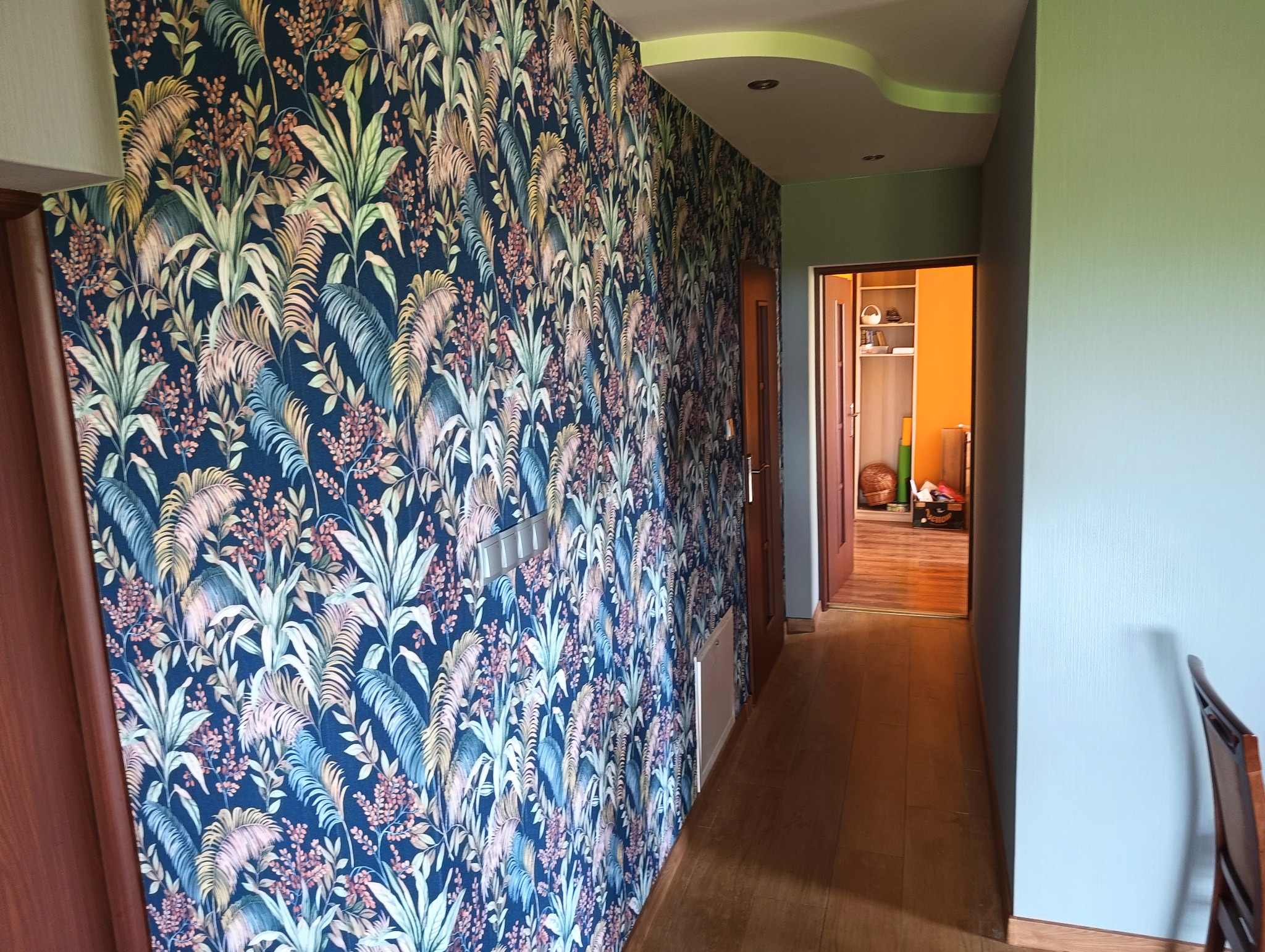 Korytarz po remoncie: ściana w tropikalne wzory, jasnoszara ściana, drewniana podłoga i drzwi do pokoju z pomarańczową ścianą w tle. Widoczne detale wykończenia.