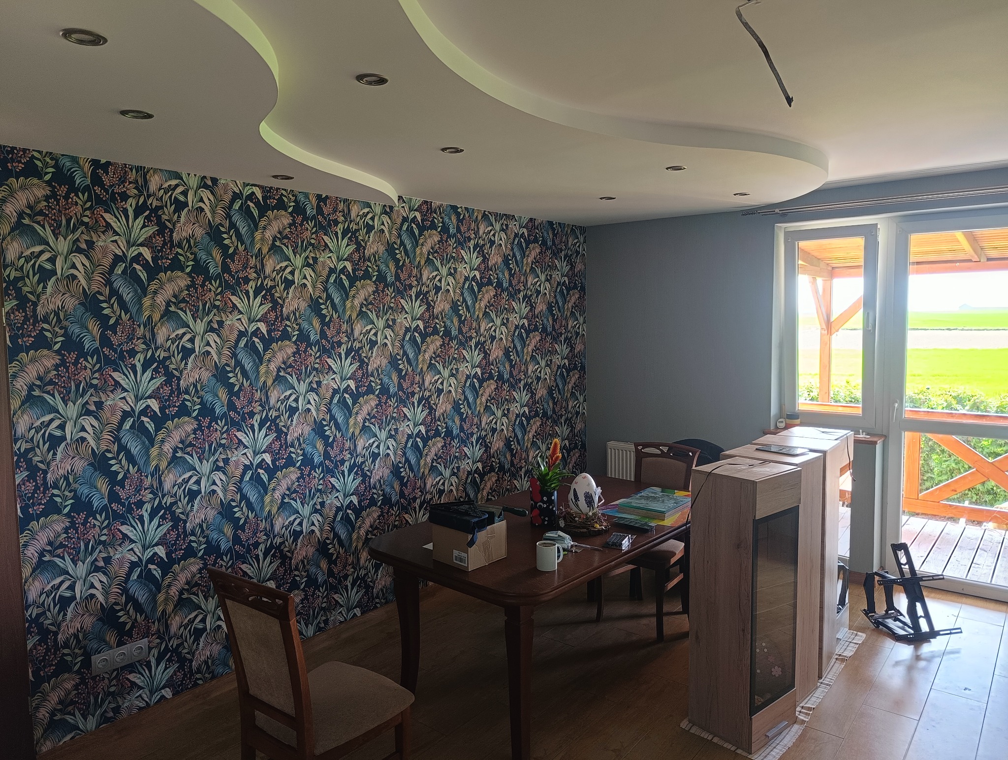Pokój po malowaniu w Szczepanowie: ściana z tapetą w roślinny wzór, szara ściana, okno z widokiem na zewnątrz, meble w pomieszczeniu. Nowoczesny design.