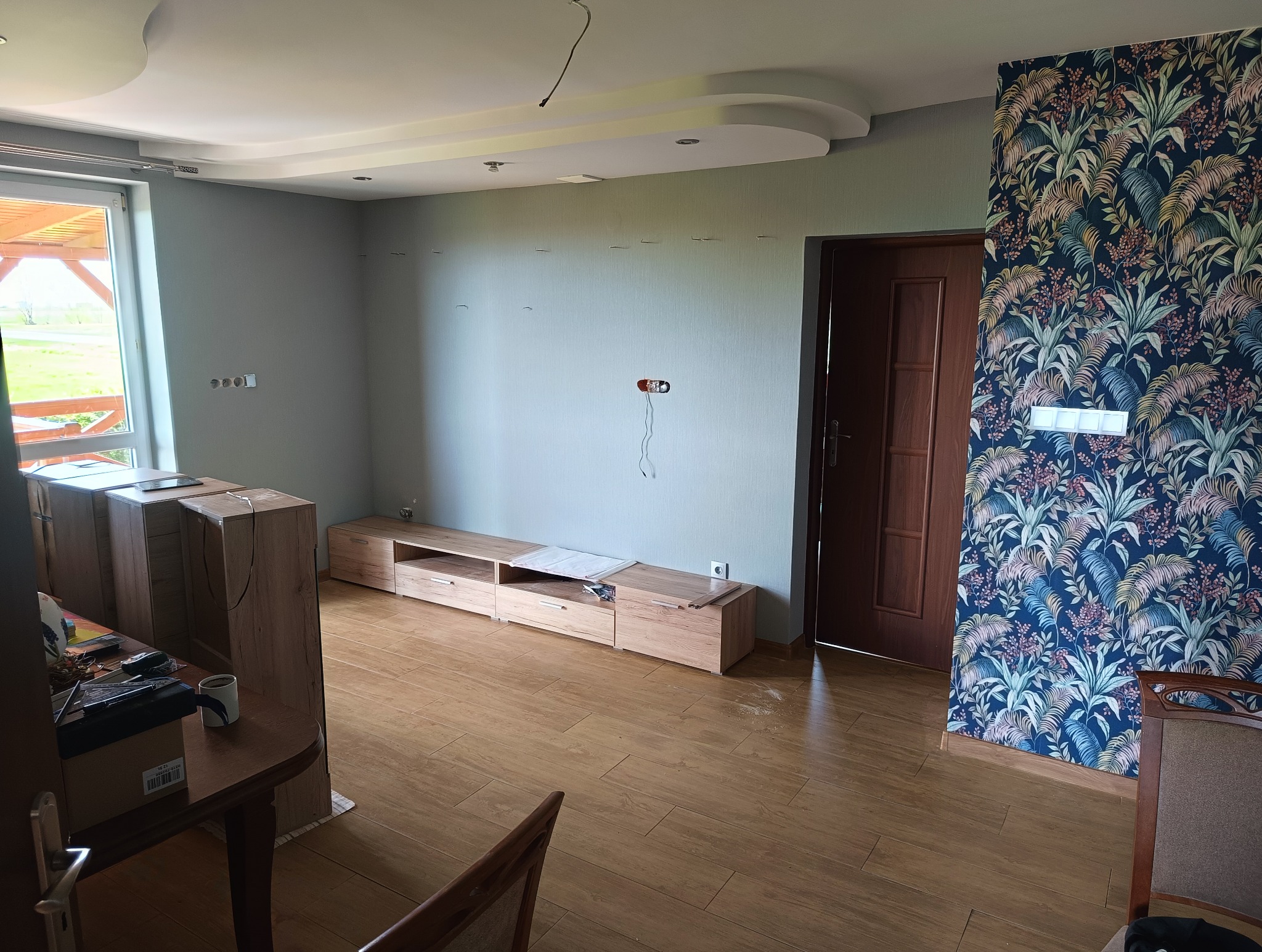 Wnętrze pokoju w trakcie wykańczania: częściowo złożona szafka RTV, kartony, wzorzysta tapeta na ścianie, drewniana podłoga. Widoczne gniazdka elektryczne.