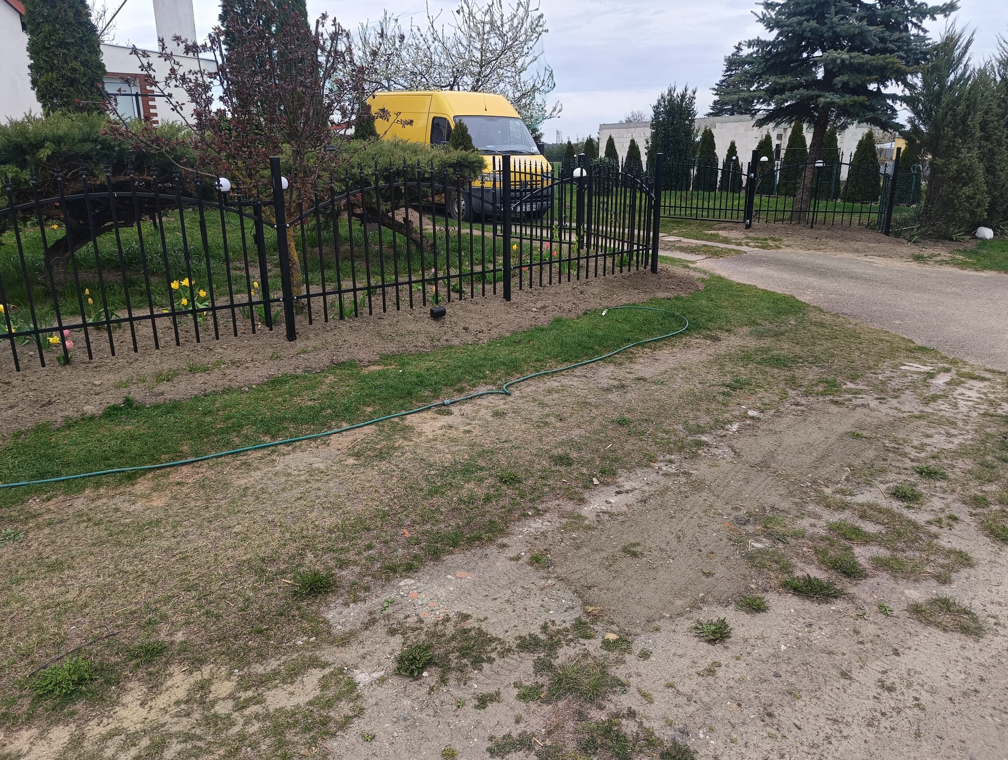 Czarny, metalowy płot panelowy z bramą wjazdową. W tle żółty samochód dostawczy i drzewa iglaste. Na pierwszym planie trawnik i fragment węża ogrodowego.
