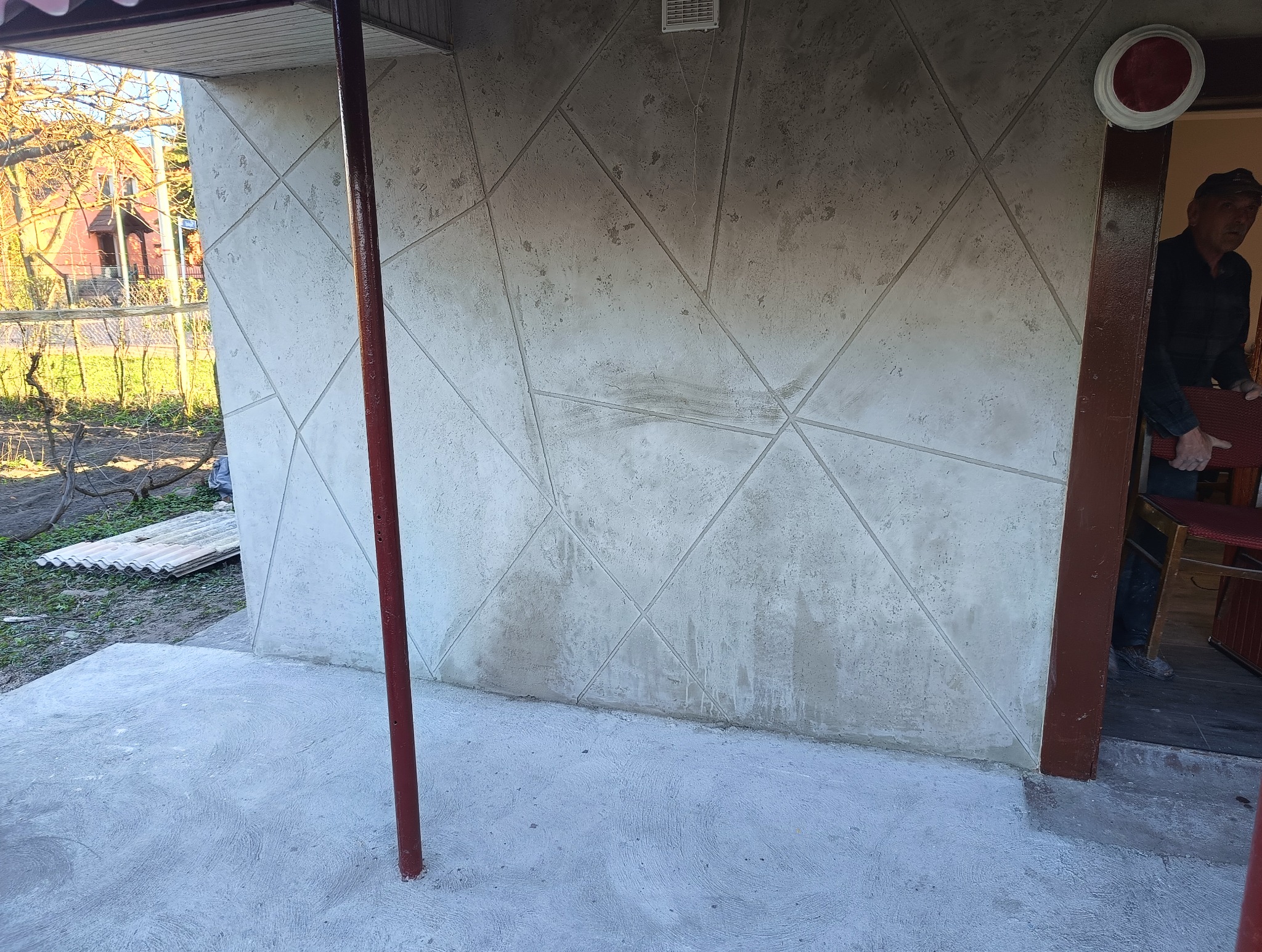 Ściana budynku z dekoracyjną, geometryczną fakturą w odcieniach szarości, widoczna część podłogi i fragment osoby w tle.