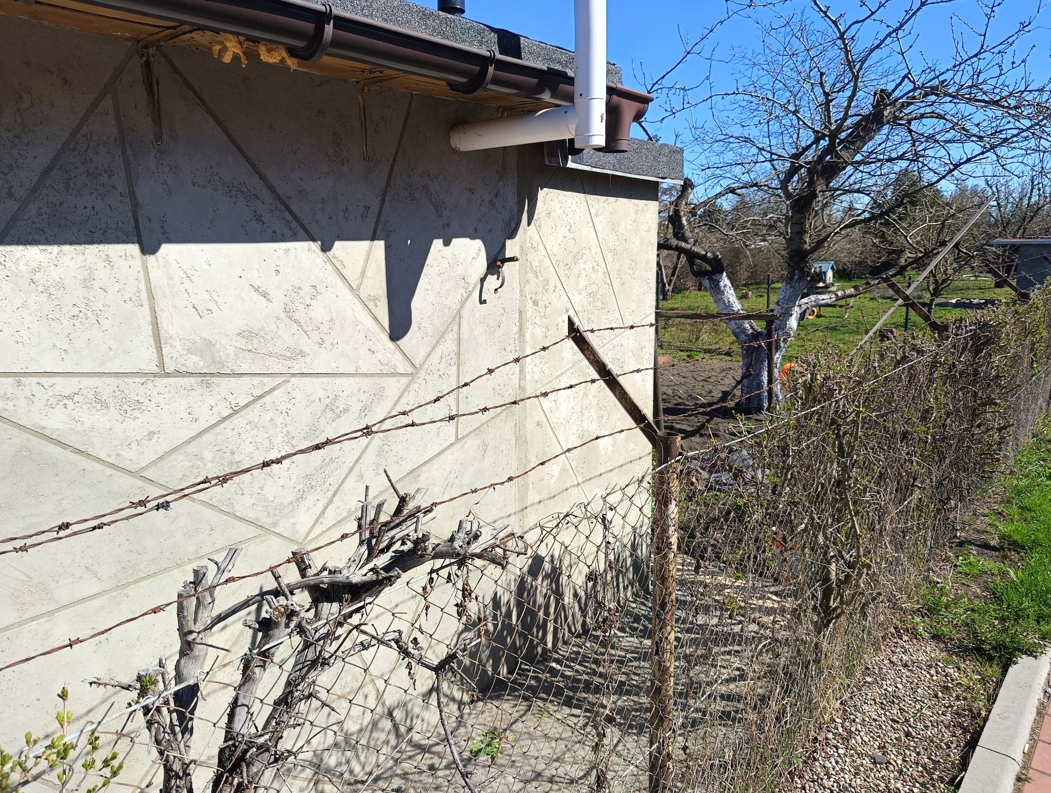 Metalowa siatka ogrodzeniowa z drutem kolczastym, przylegająca do ściany budynku z brązową rynną. W tle ogród z drzewami i niebieskim niebem.