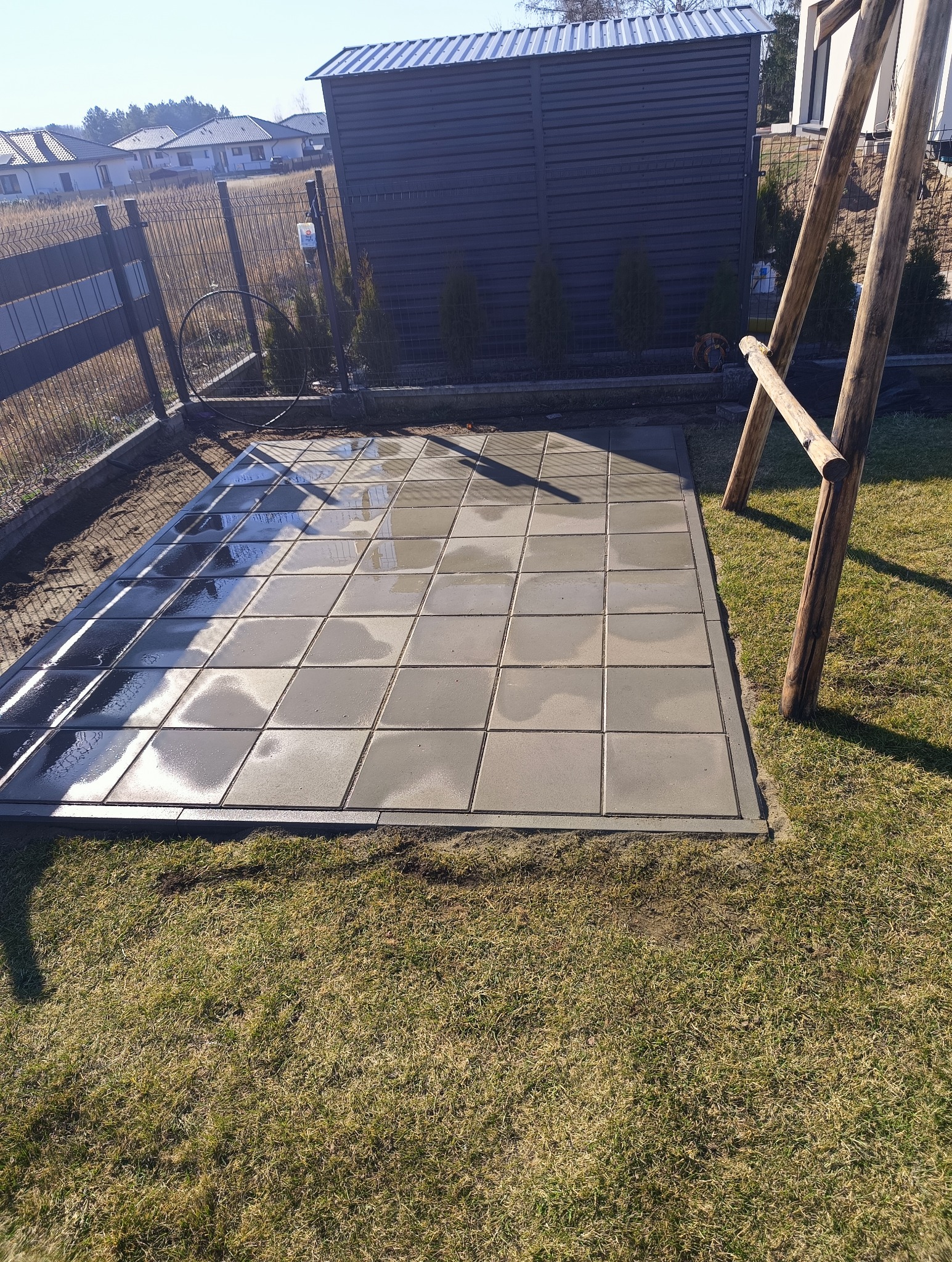 Świeżo ułożona, mokra kostka brukowa na posesji, z widocznym fragmentem ogrodzenia i drewnianej konstrukcji huśtawki w tle, w słoneczny dzień.