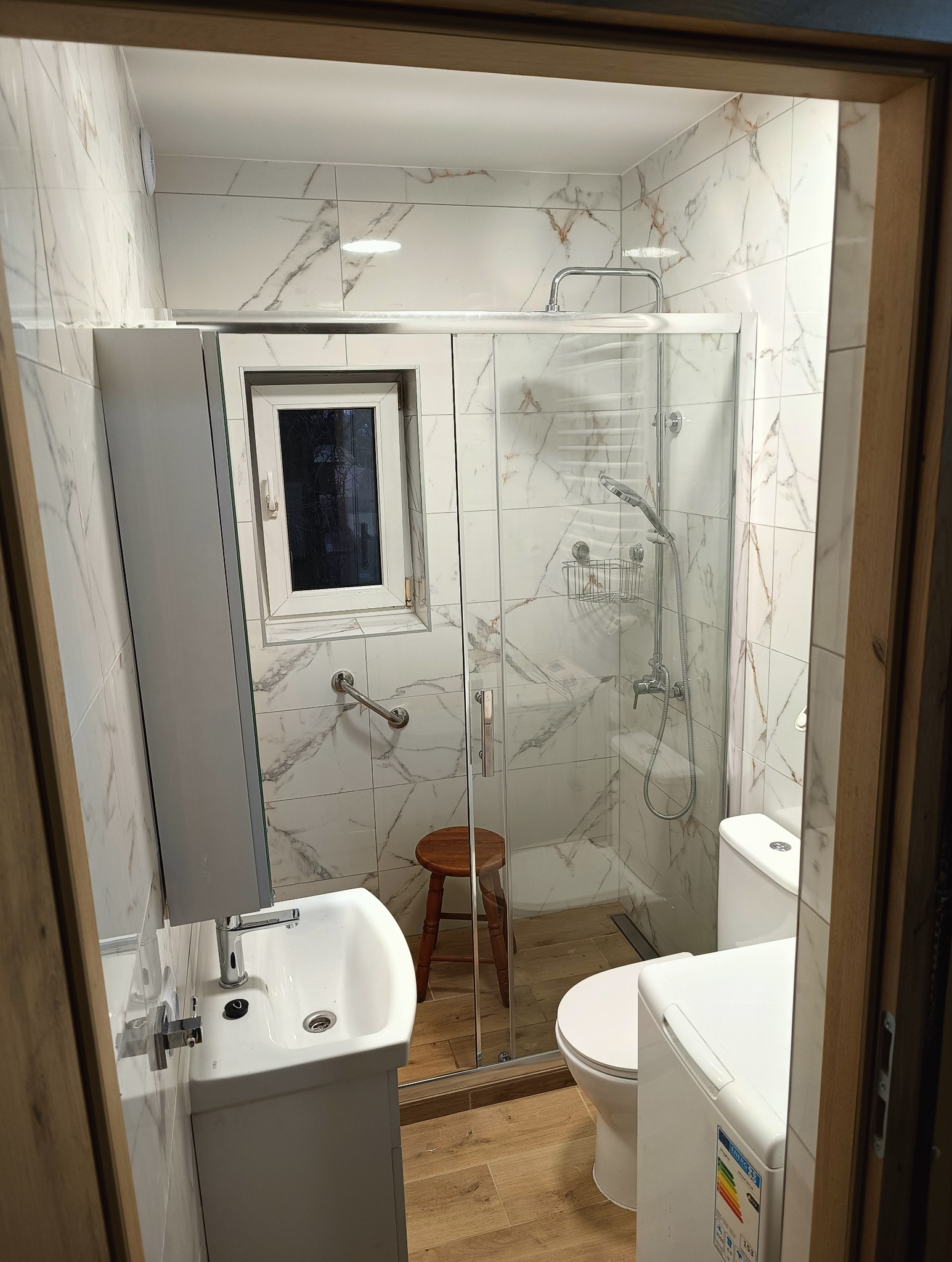 Wykończona łazienka z marmurowymi płytkami, kabiną prysznicową ze szklanymi drzwiami, umywalką z szafką i drewnianym stołkiem. Widoczne udogodnienia dla osób z niepełnosprawnościami.