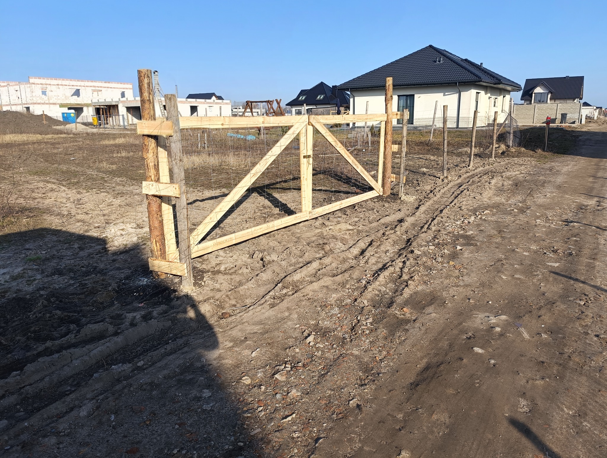 Surowa, drewniana brama z siatką w ogrodzeniu na tle budynków w stanie surowym i gotowych domów. Ślady opon na ziemi. Ujęcie w słoneczny dzień.