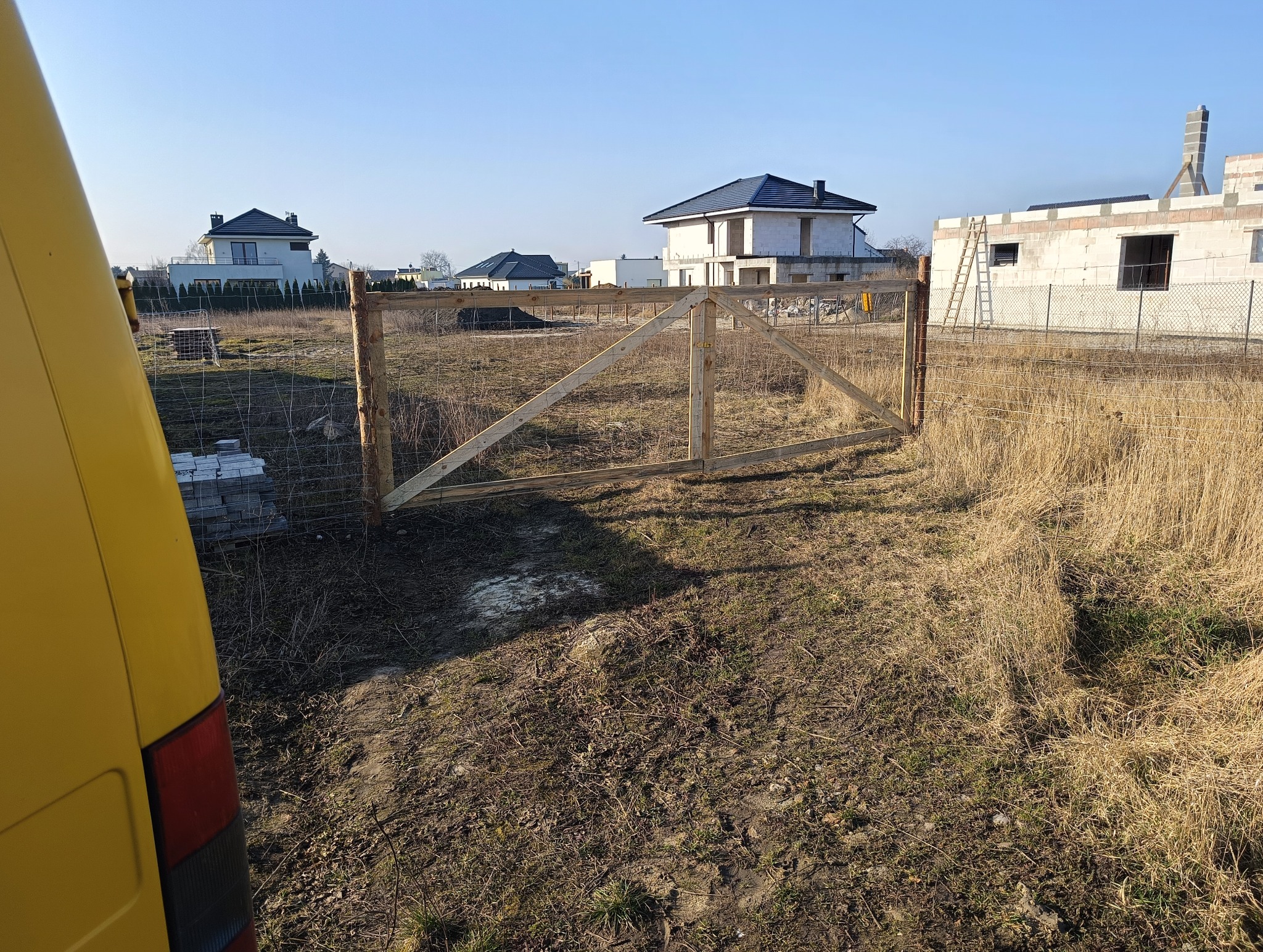 Drewniana brama z siatką na działce budowlanej w słoneczny dzień. Część żółtego samochodu dostawczego po lewej. Domy w tle. Widoczny etap budowy.