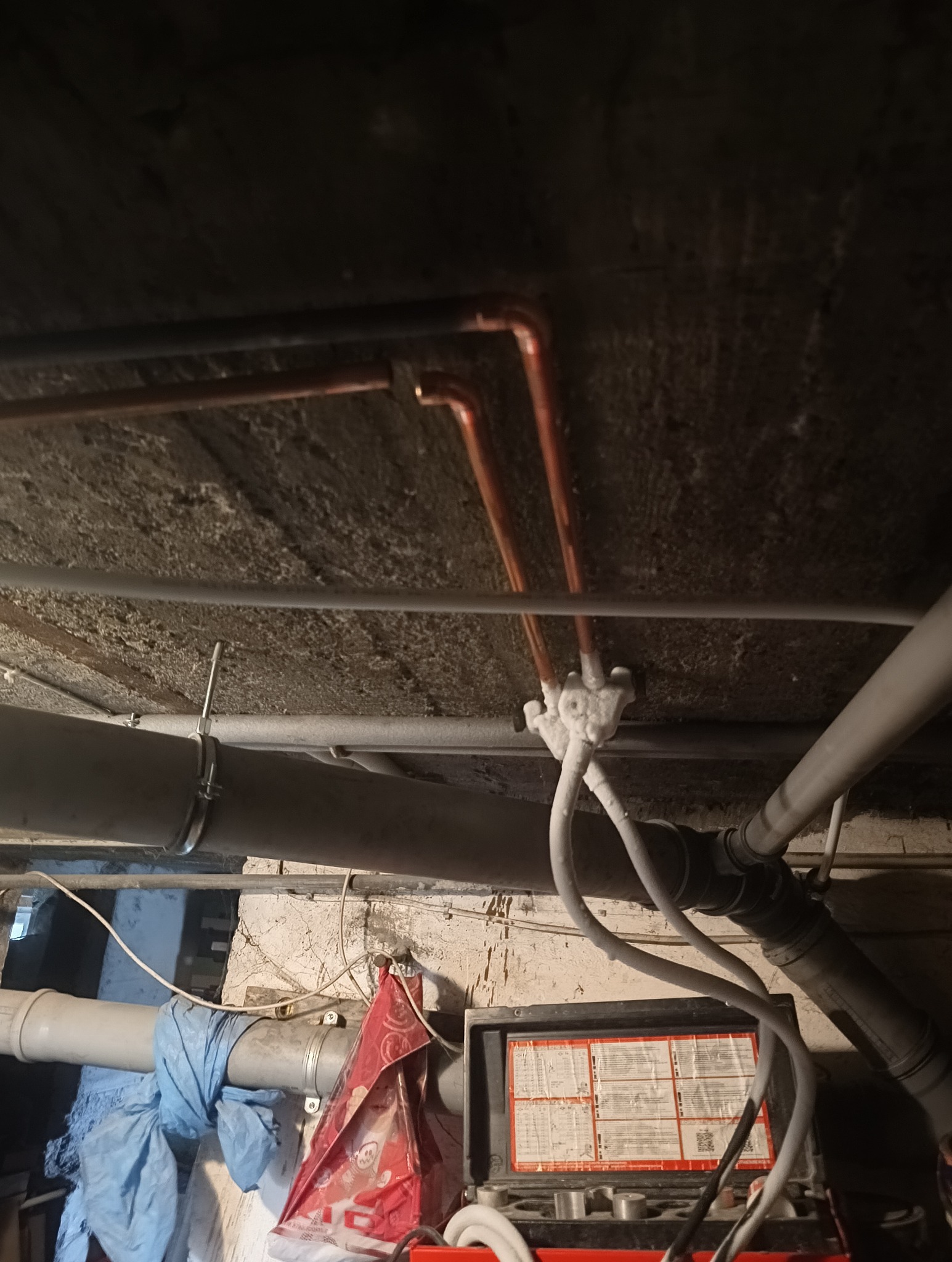 Instalacja rur miedzianych i PCV pod stropem, widoczne łączenia i mocowania. Ujęcie od dołu, ciemne tło uwydatnia detale instalacji.