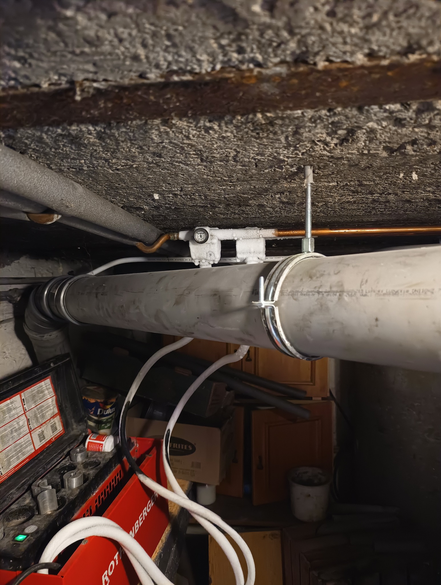 Szara rura kanalizacyjna pod sufitem, przymocowana metalowymi obejmami, z widocznym manometrem i urządzeniem Rothenberger. Instalacja w piwnicy lub innym pomieszczeniu gospodarczym.