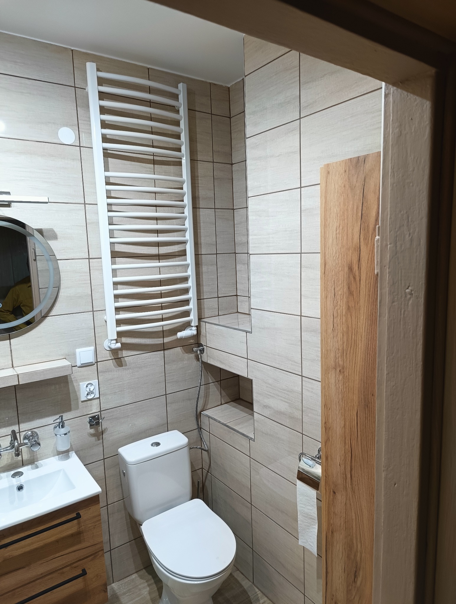 Nowoczesna łazienka z beżowymi płytkami, białym grzejnikiem drabinkowym i okrągłym lustrem z podświetleniem. Widoczna toaleta i umywalka z szafką w kolorze drewna.