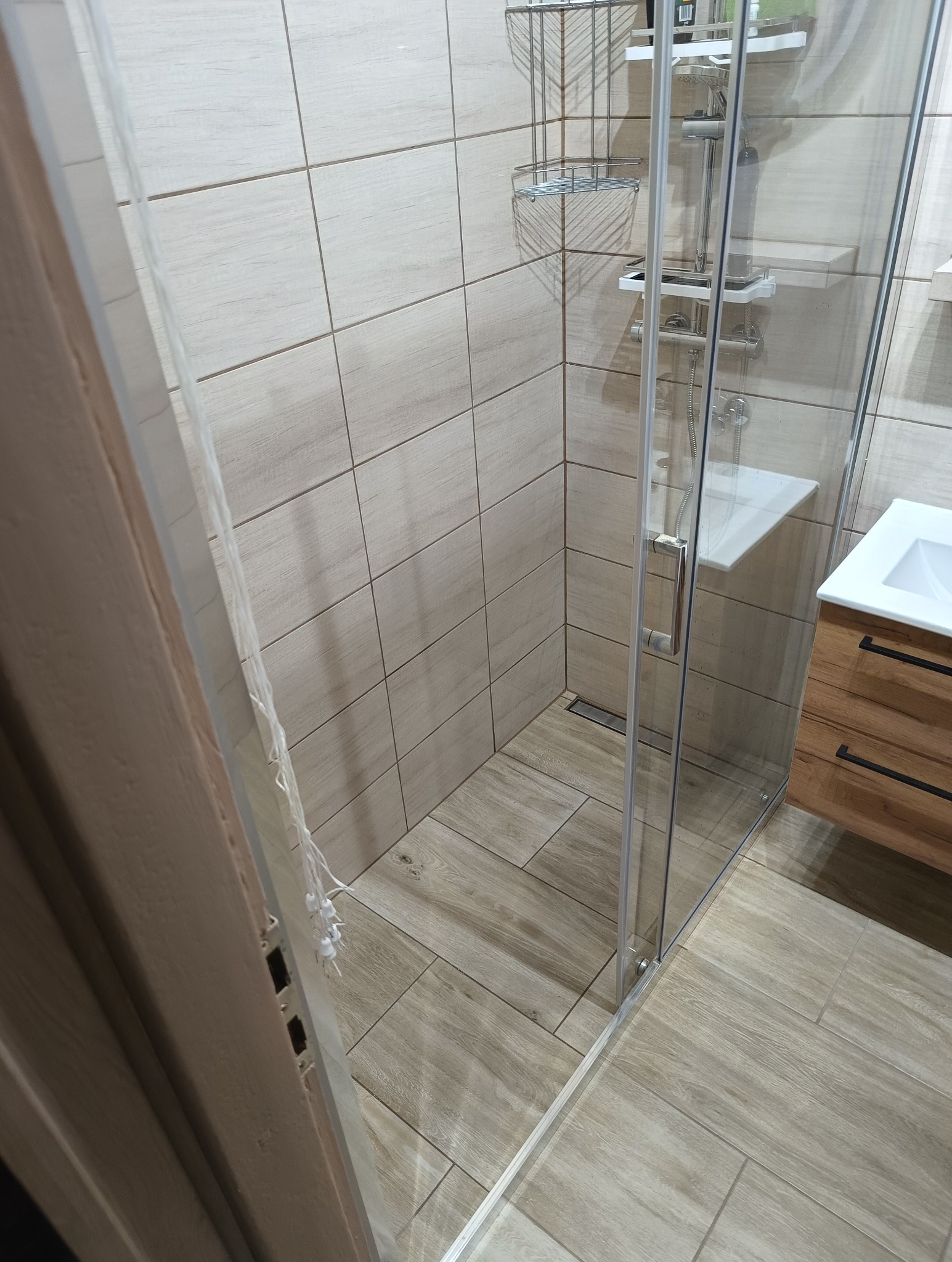 Nowoczesna kabina prysznicowa z drewnopodobnymi płytkami i szklaną ścianą, chromowane akcenty, widok na umywalkę z szafką. Minimalistyczny design.