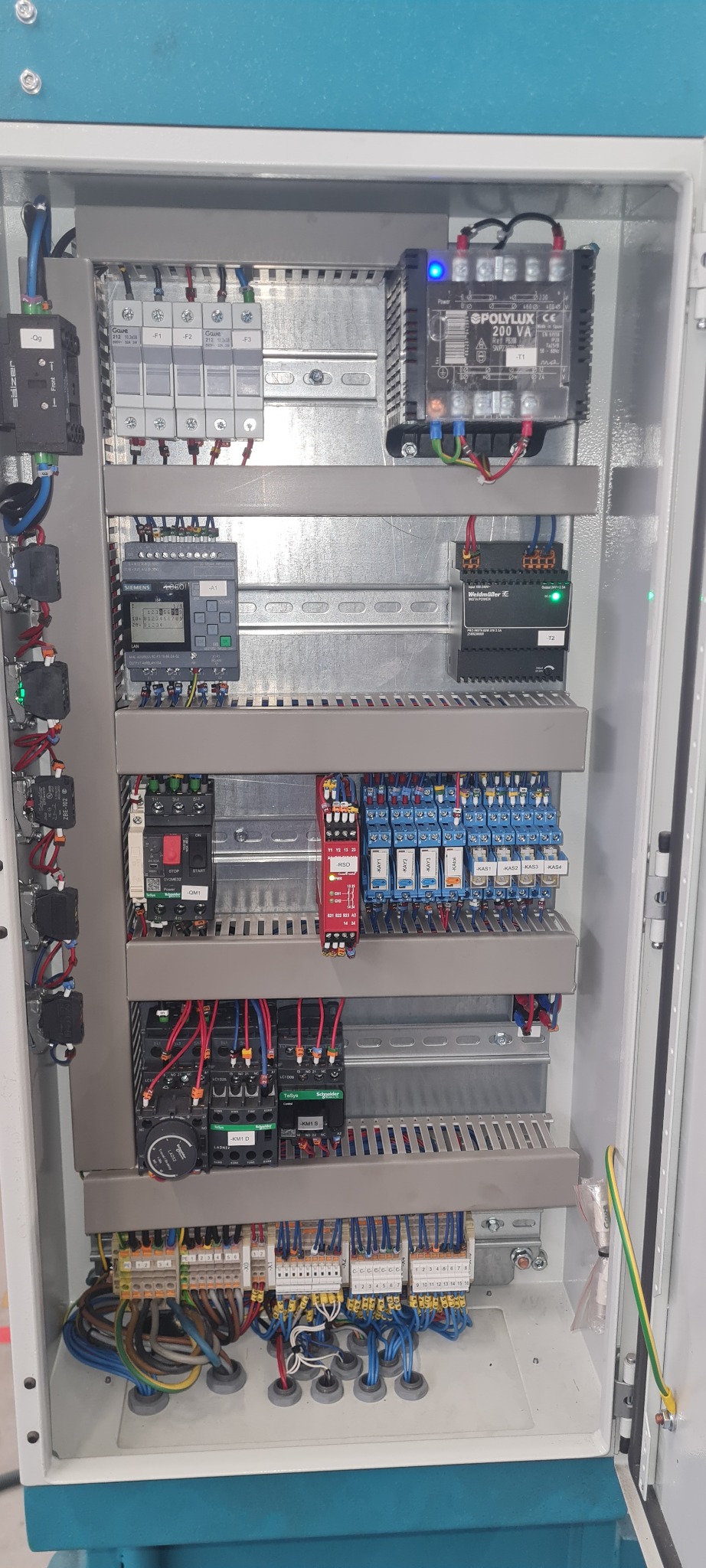Otwarta szafa sterownicza z widocznymi elementami instalacji elektrycznej: bezpieczniki, przekaźniki, sterownik PLC Siemens, zasilacz Weidmuller i listwy zaciskowe z okablowaniem.