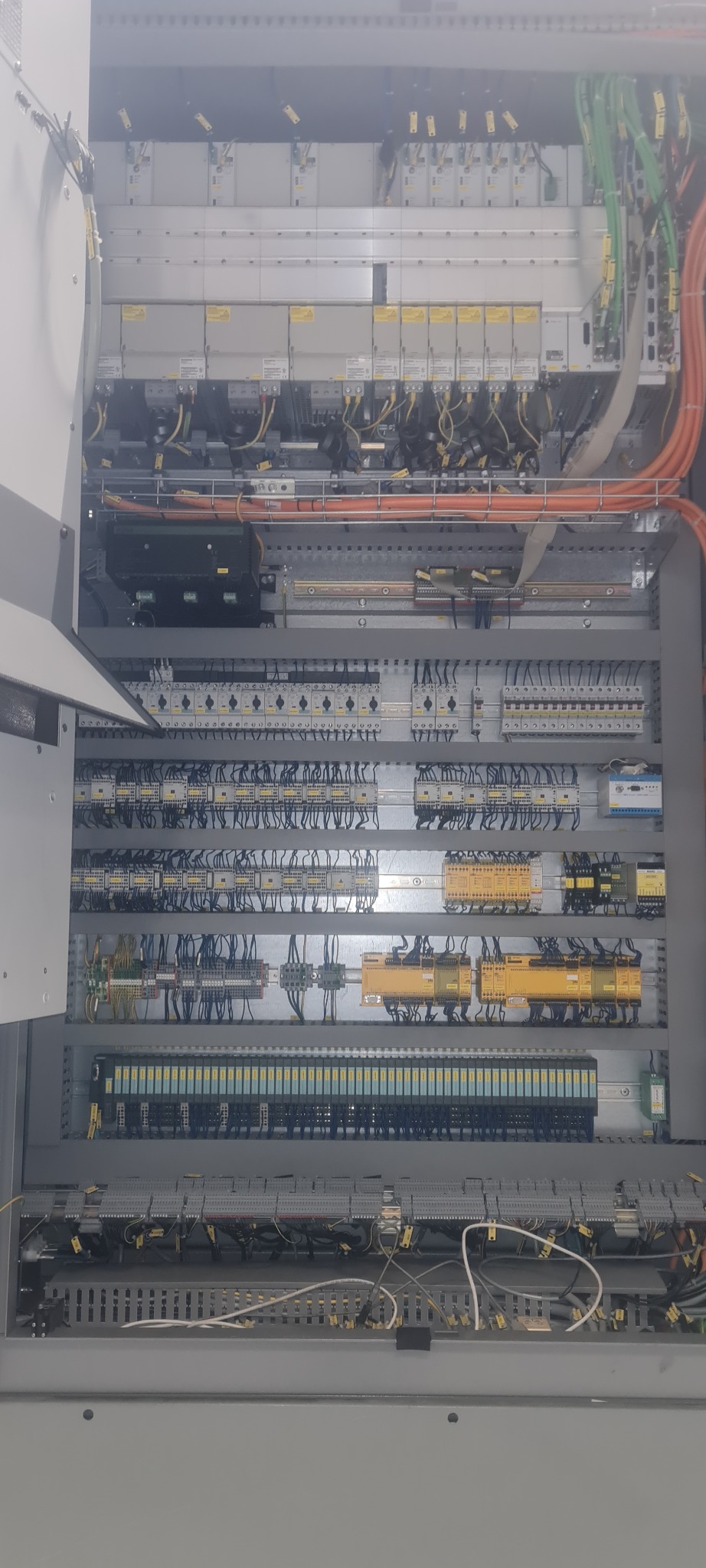Otwarta szafa sterownicza z widocznymi modułami PLC, bezpiecznikami, przekaźnikami i okablowaniem w kolorach pomarańczowym i zielonym.